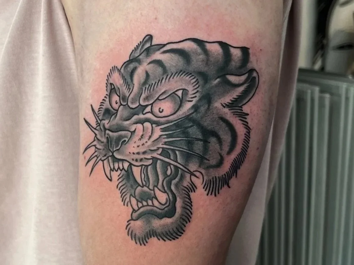 Tattoo of a fierce tiger's face on upper arm, featuring bold black outlines and shading.