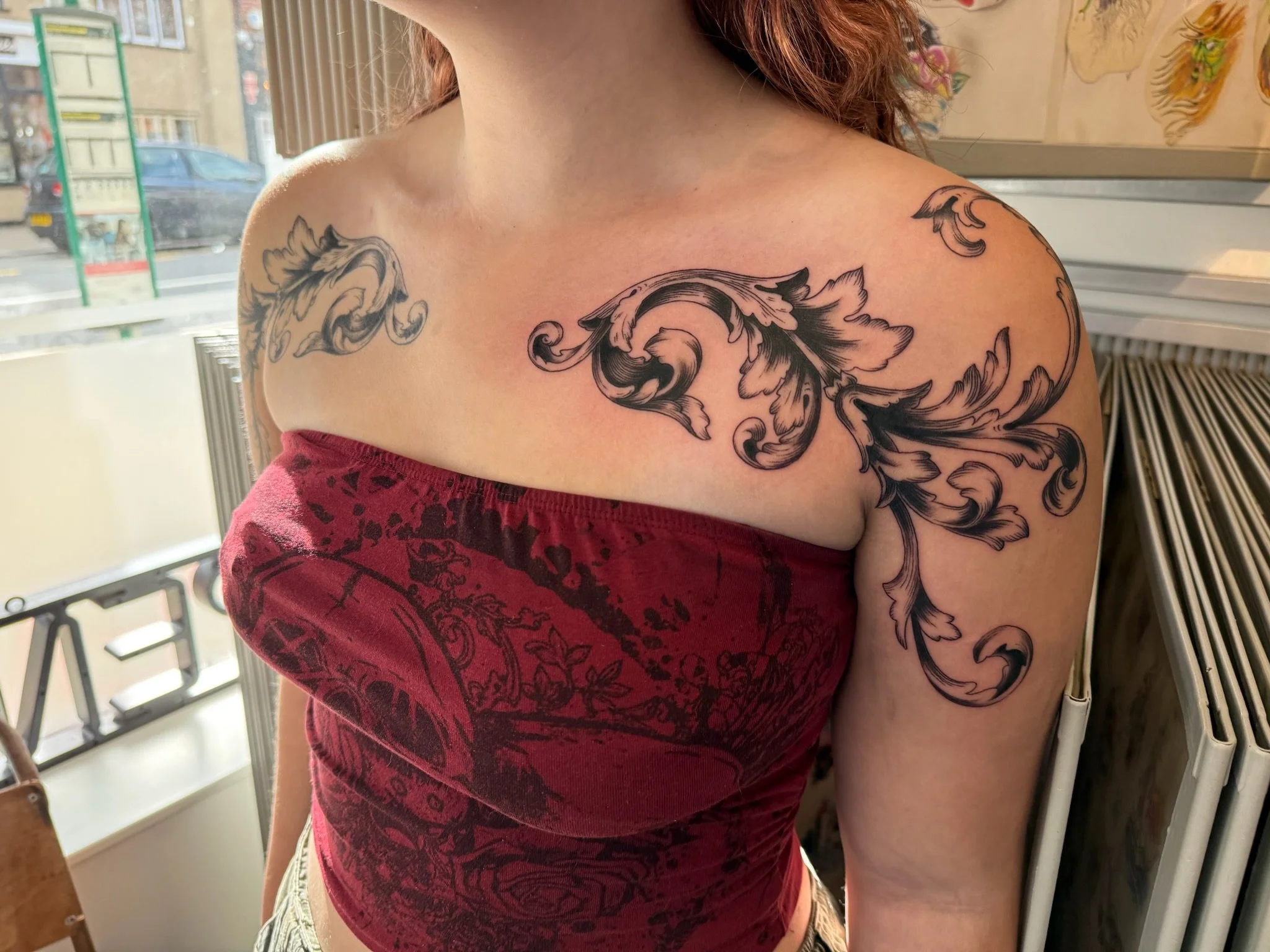 Close-up of a woman's upper body showing a shoulder and arm tattoo with intricate, flowing floral and scrollwork patterns. The woman is wearing a red shirt and has red hair, with a background of a window and a railing.