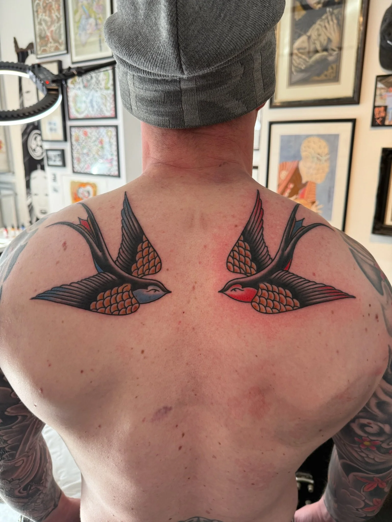Back of person with a tattoo of two koi fish on the upper back, wearing a gray beanie, exposed shoulders, visible tattoos on arms, in an art gallery with framed artworks on the wall.