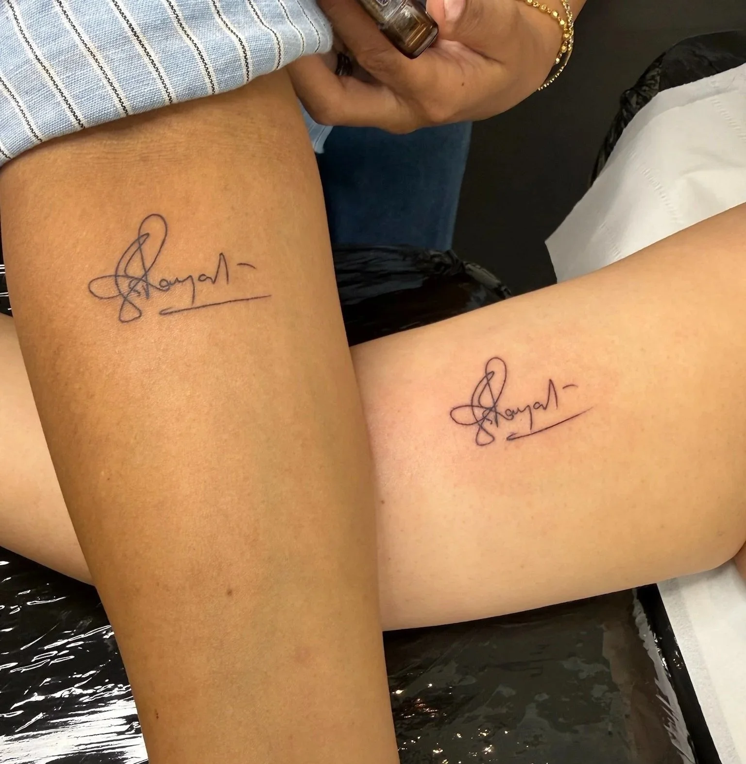 Two people with matching tattoo signatures on their thighs, one person is wearing black shorts and the other person is wearing a striped shirt, sitting on a surface covered with black plastic.