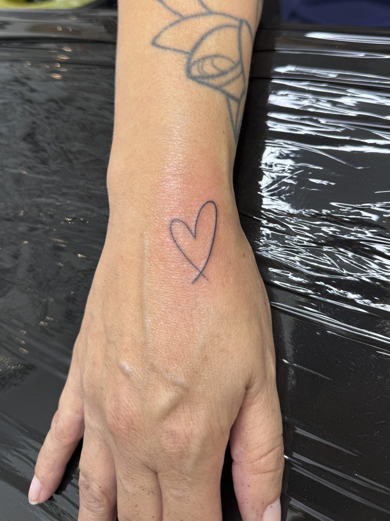 A person's hand with a simple outline heart tattoo on the wrist, resting on a black surface with a tattooed arm partially visible in the background.