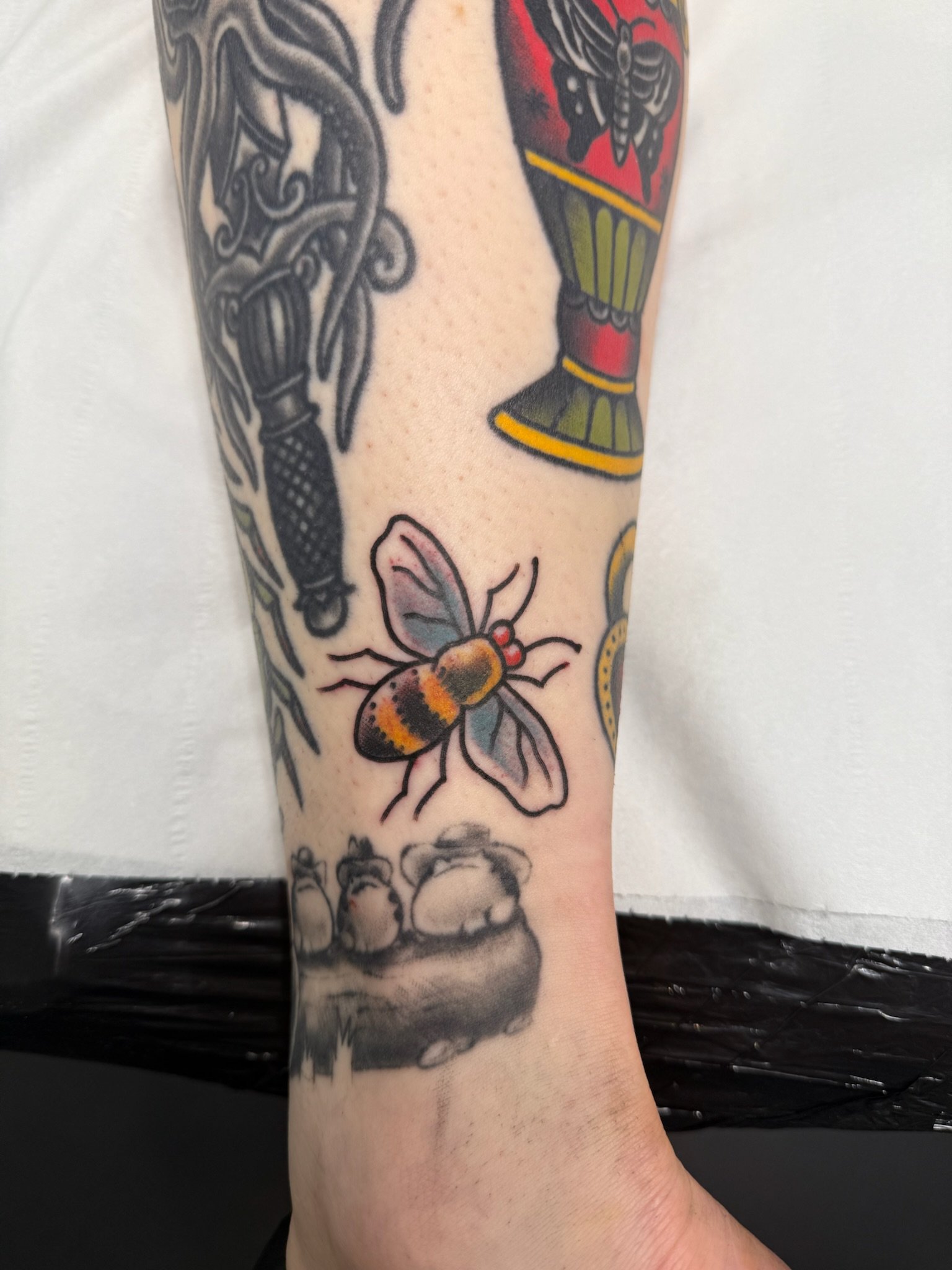 Tattoo of a bee with colorful wings on a person's arm with other tattoo designs visible.