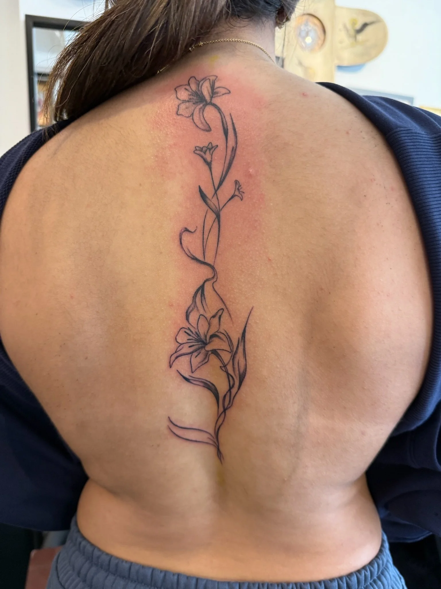 A person's shoulder and upper back featuring a black outline tattoo of three lilies with long stems, one large flower at the top, and two smaller flowers in the middle and bottom.