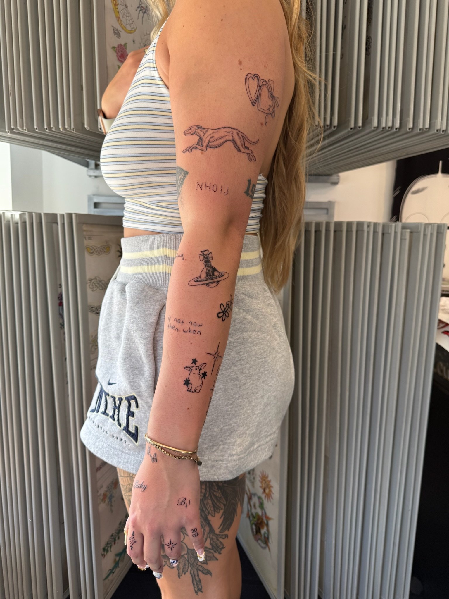 A woman with several tattoos on her arm, hand, and thigh, wearing a sleeveless striped top and gray shorts, standing in front of a metallic partition and decorative wallpaper.