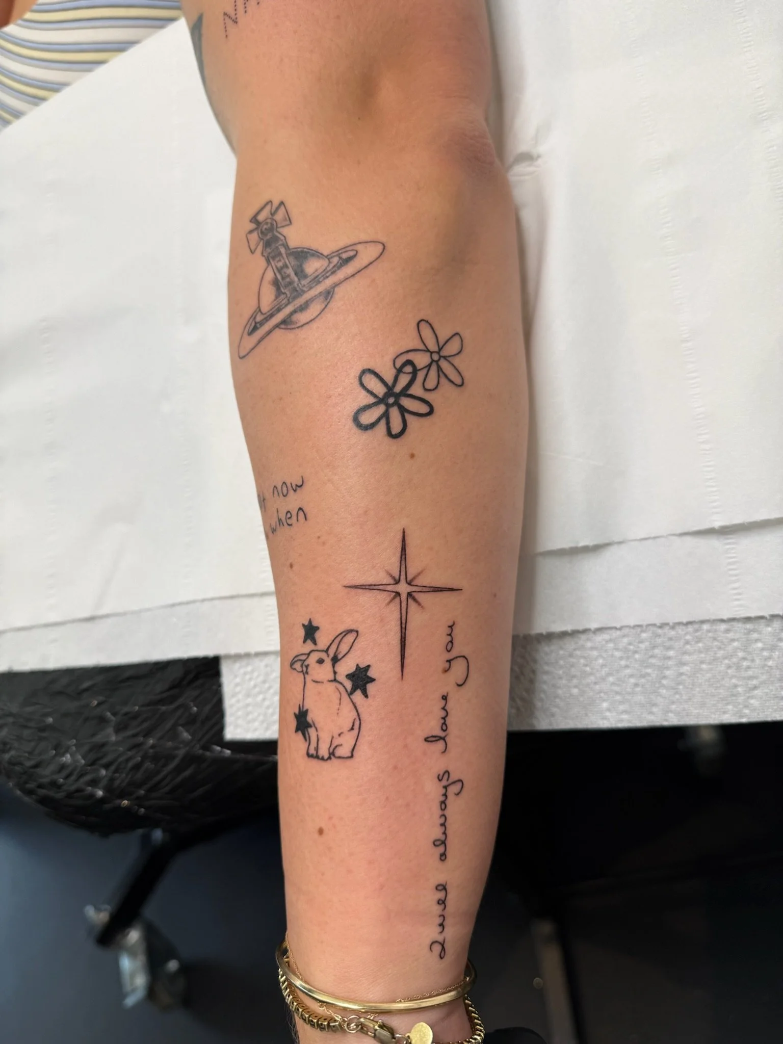 A person's forearm with multiple tattoos, including a planet with rings and a small creature, outlined stars, and cursive writing.