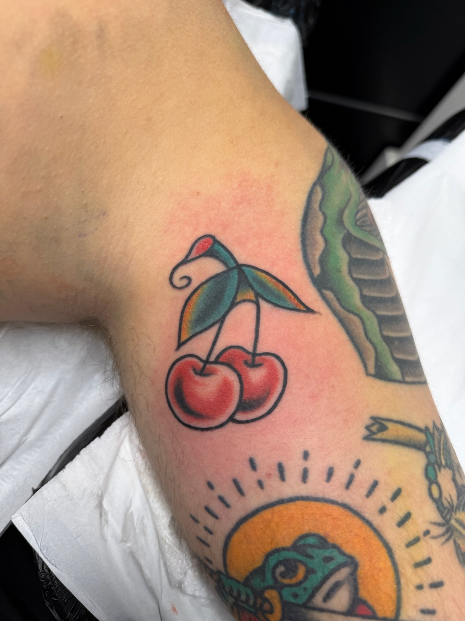 Close-up of a person's upper arm with a colorful tattoo of cherries and a green leaf, and part of a tattoo of a sun with rays.