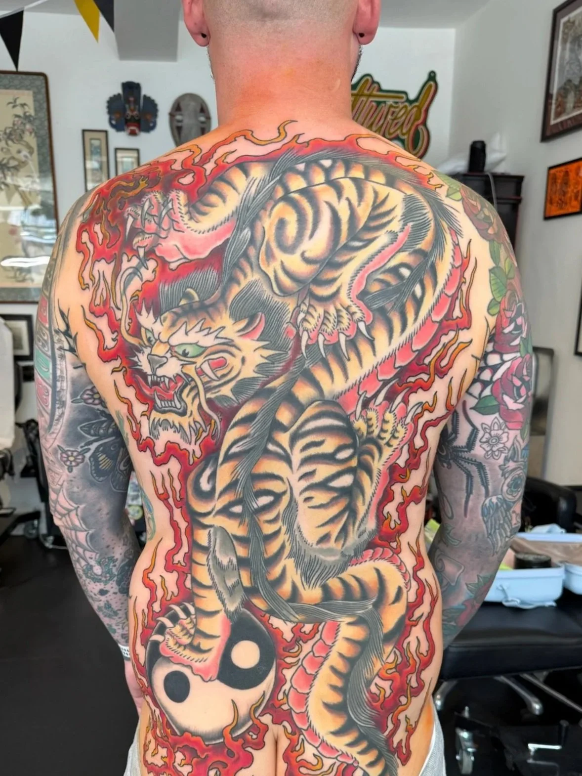 Back tattoo of a fierce tiger with flames and red accents, tattooed on a person's back in a tattoo studio.