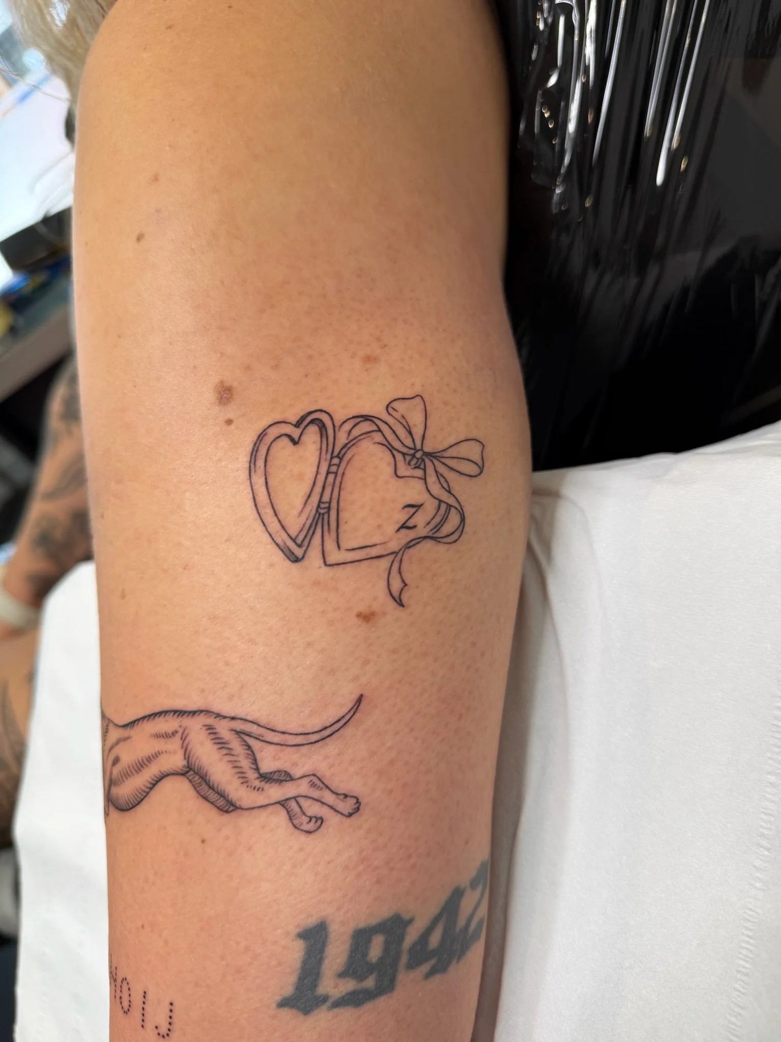 Tattoo of a heart and bow on upper arm, partially visible styles and text on lower arm.