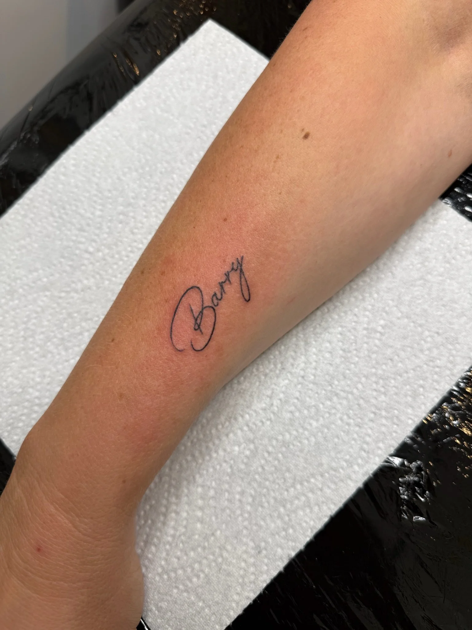 A forearm with a small mole and a tattoo that reads 'Bay' is resting on a white paper towel, on a black surface.