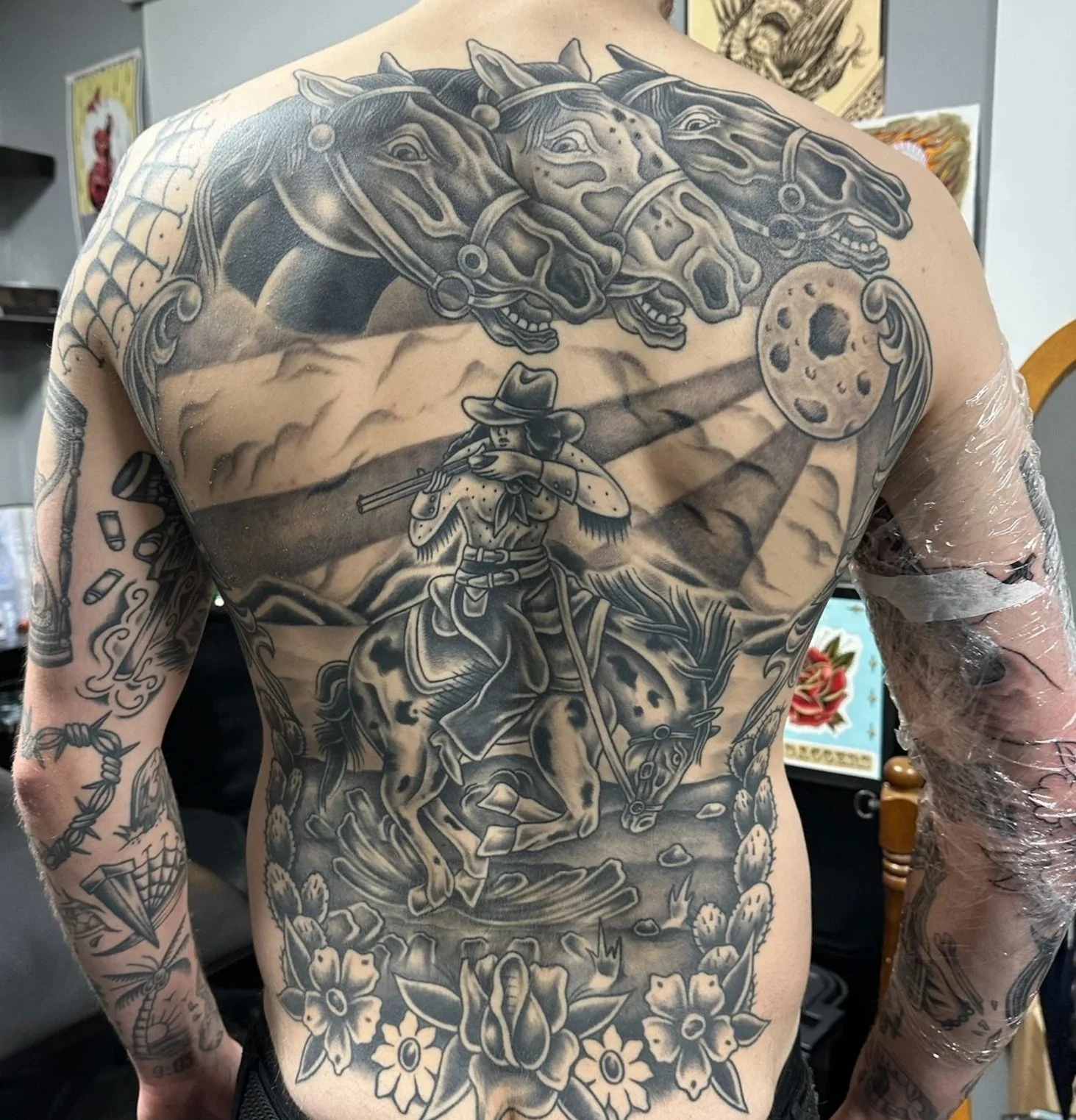 Large black and gray tattoo on a man's back features three horse heads at the top, a woman in a wide-brimmed hat with a rifle in the middle, riding a horse among cacti and rocks, with a moon in the background. Additional tattoos are visible on his arms.