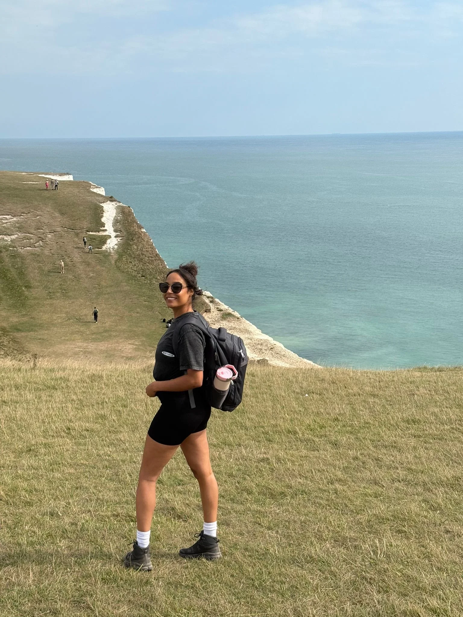 A woman with sunglasses and a backpack standing on a grassy hill near the coast, with a blue sea and cliffs in the background.
