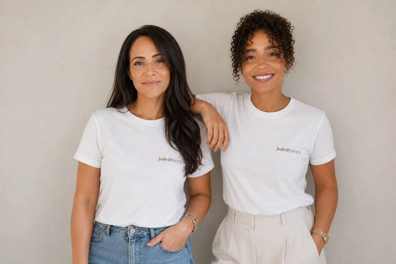 Two women standing side by side against a plain background, smiling. Both women are wearing white t-shirts with the text 'Jade&Jorgie'. One woman has long dark hair and is wearing blue jeans, while the other has curly hair and is wearing light-colored pants. The woman with curly hair has her arm resting on the other woman's shoulder.