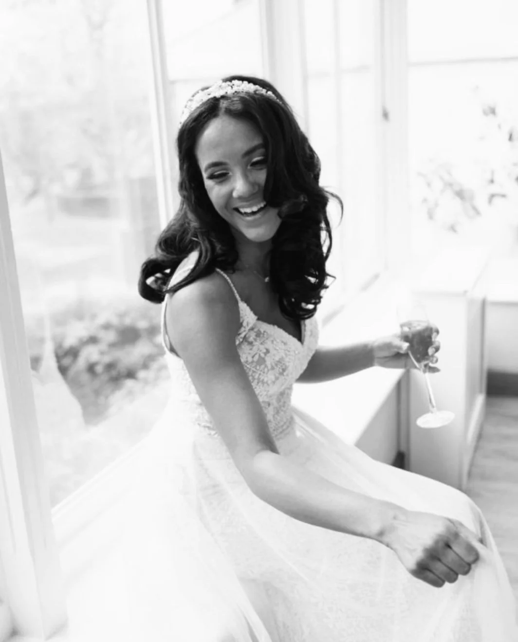 Black and white photo of a young woman in a wedding dress sitting by a window, smiling, holding a glass of champagne.