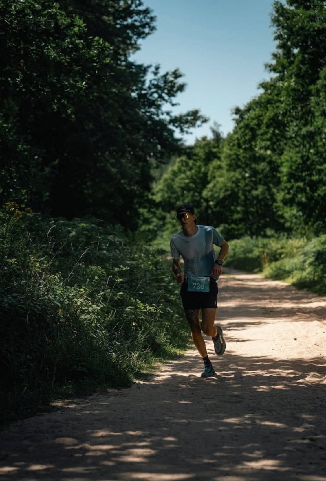 A person running on a dirt trail surrounded by lush green trees under a clear blue sky.
