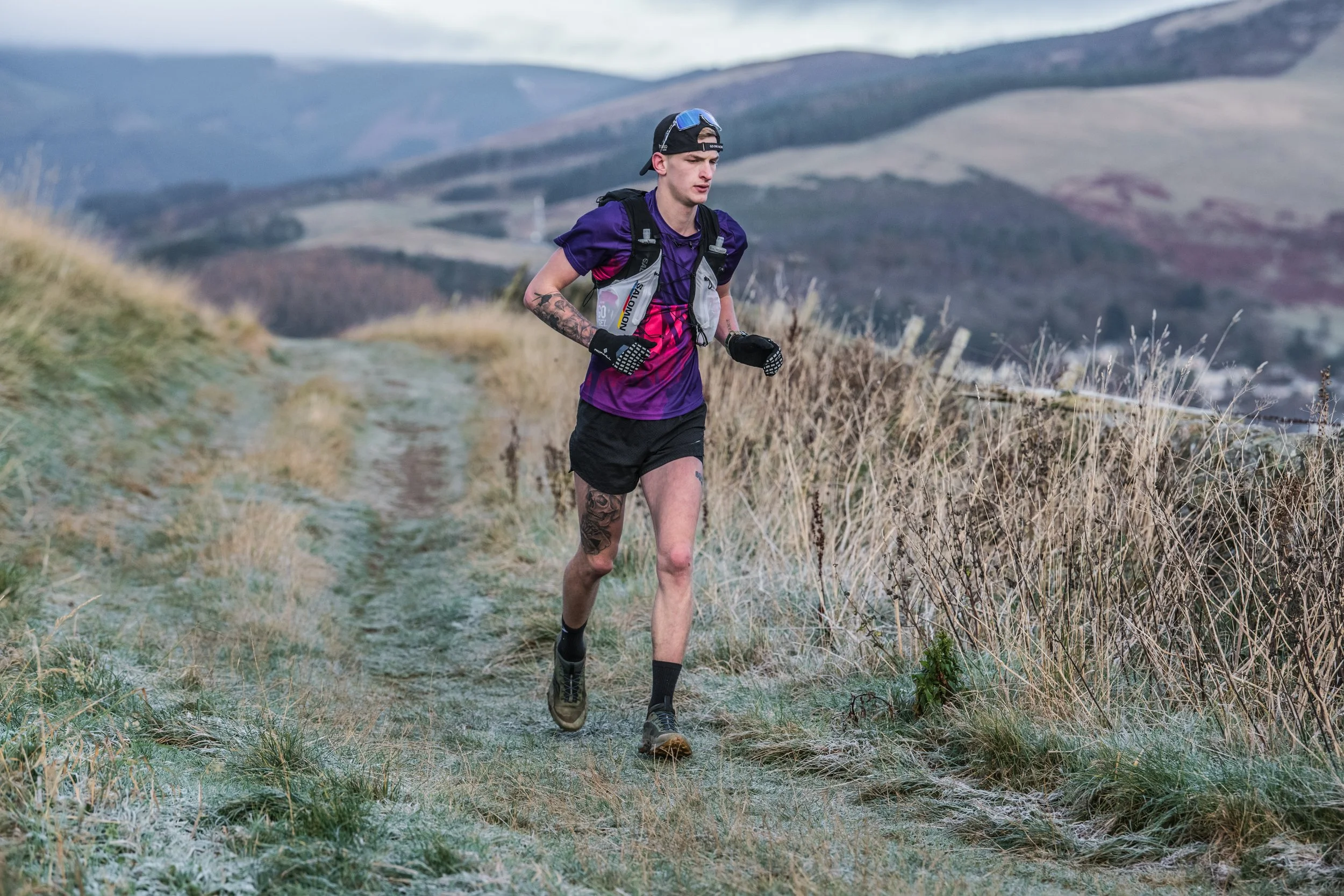 A young man running on a trail in a mountainous area, wearing athletic gear, a purple shirt, black shorts, and a backpack.