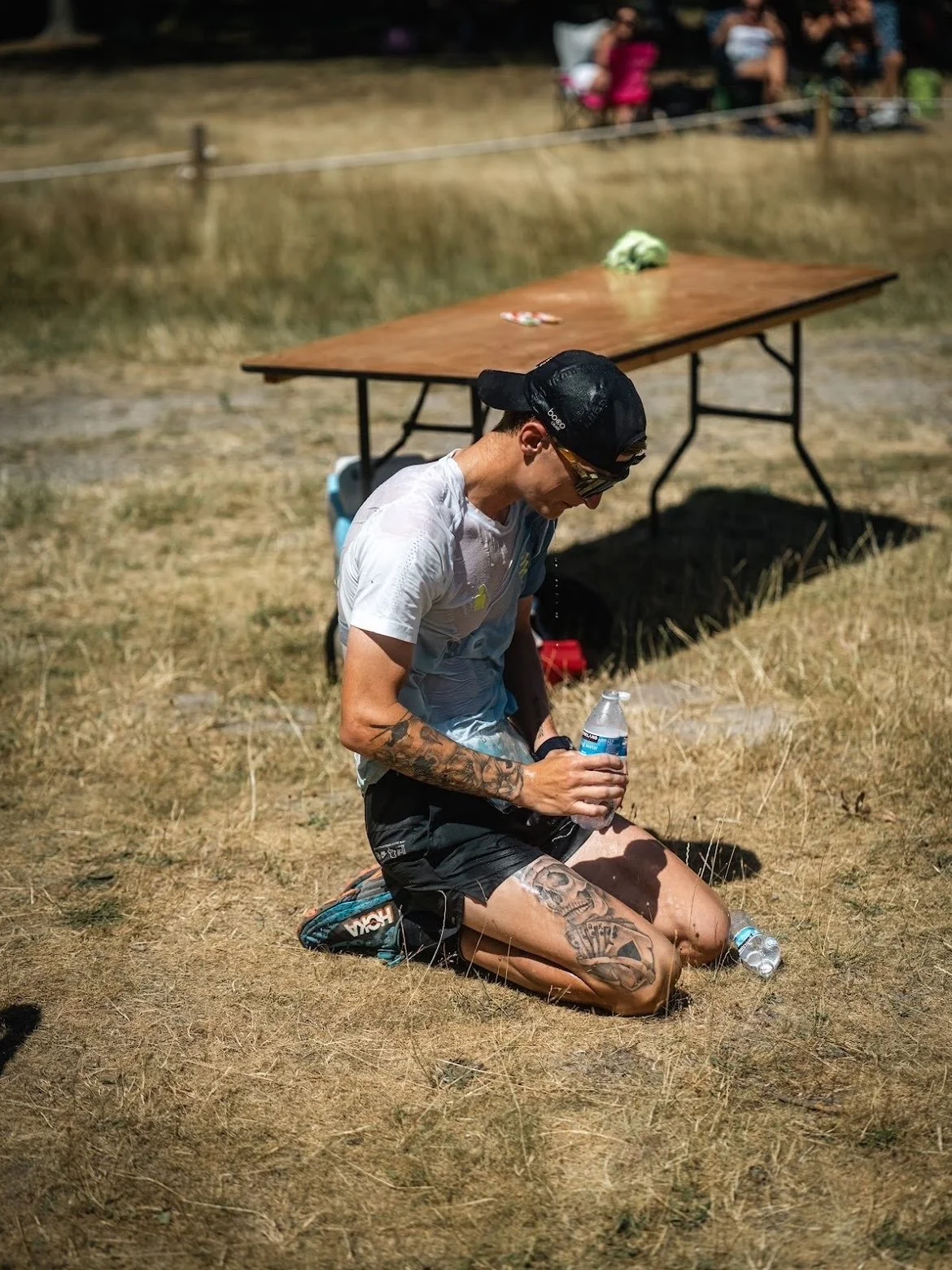 A man kneeling on dry grass holding a water bottle after a race or outdoor event, with a table and spectators in the background.