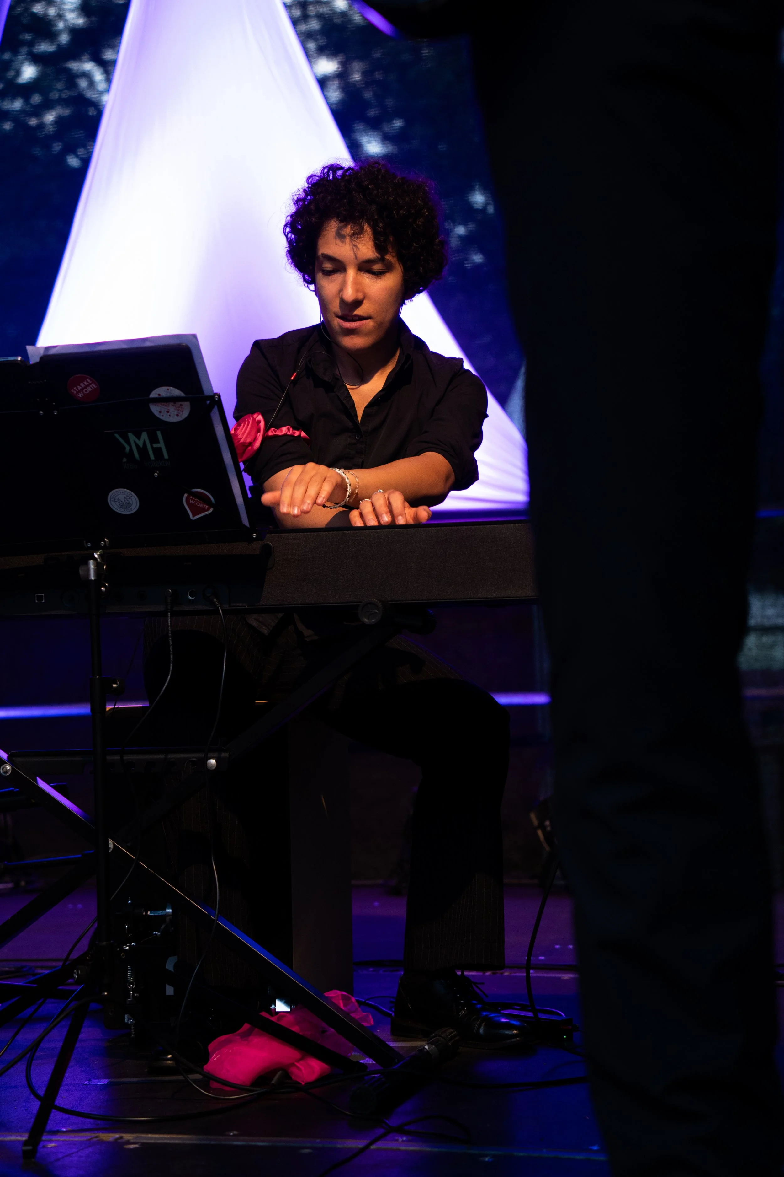 Junge Frau mit dunklen, lockigen Haaren spielt im abgedunkelten Raum an einem Keyboard, im Hintergrund eine leuchtende, rosa-farbene Lichtskulptur.