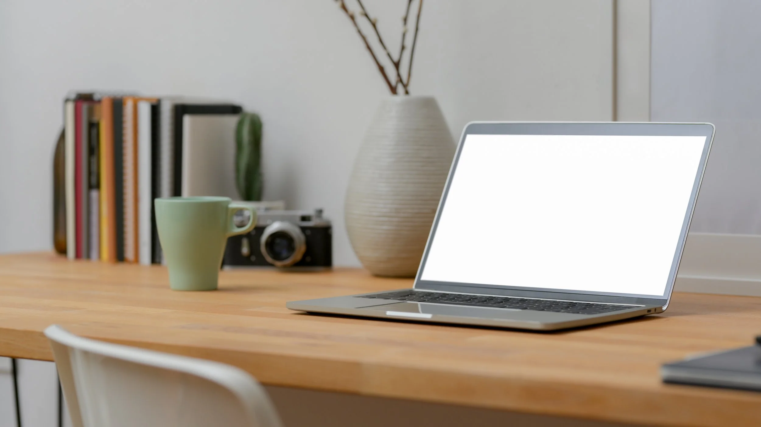 Ein Arbeitsplatz mit einem Laptop, einem grünen Kaffeebecher, einem Buchregal, einer Kamera, einer Vase mit Zweigen und einem weißen Stuhl.