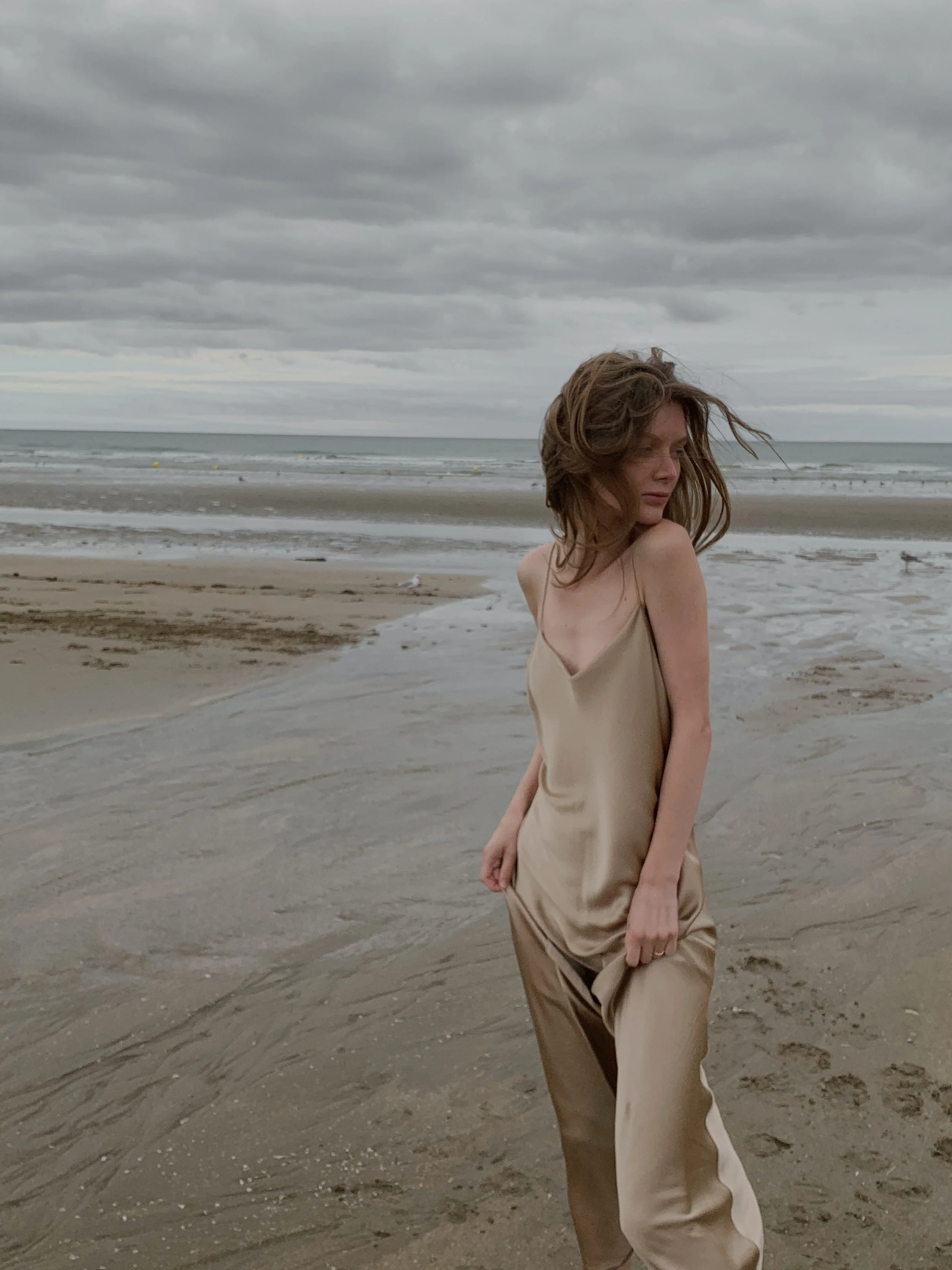 A woman with wavy brown hair standing on a beach with cloudy skies, wearing beige clothing, looking to her right.