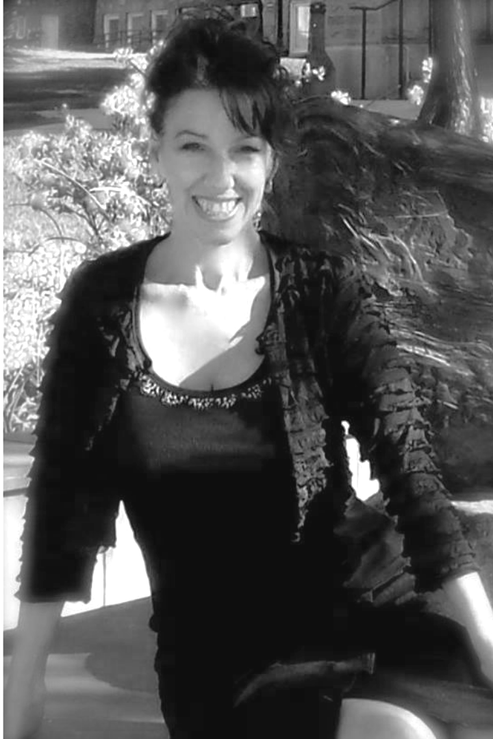 A woman outside smiling with wind-blown hair, wearing a dark top and jacket, with trees and buildings in the background.