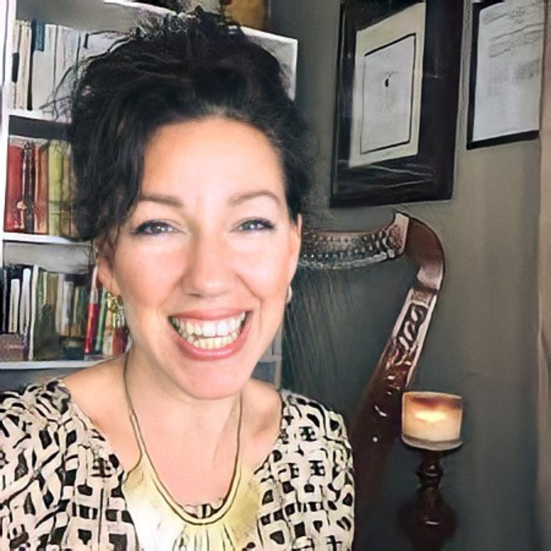 Smiling woman with short curly dark hair, wearing a patterned top and gold jewelry, in a room with bookshelves and framed pictures on the wall.