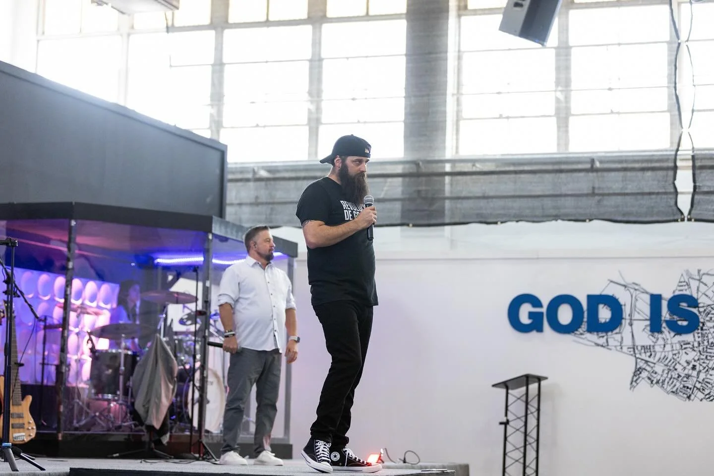 Speaker holding a microphone in front of the God Is Here backdrop.