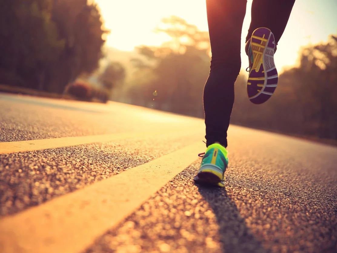Person running on a road during sunset, focusing on their shoes and lower legs.