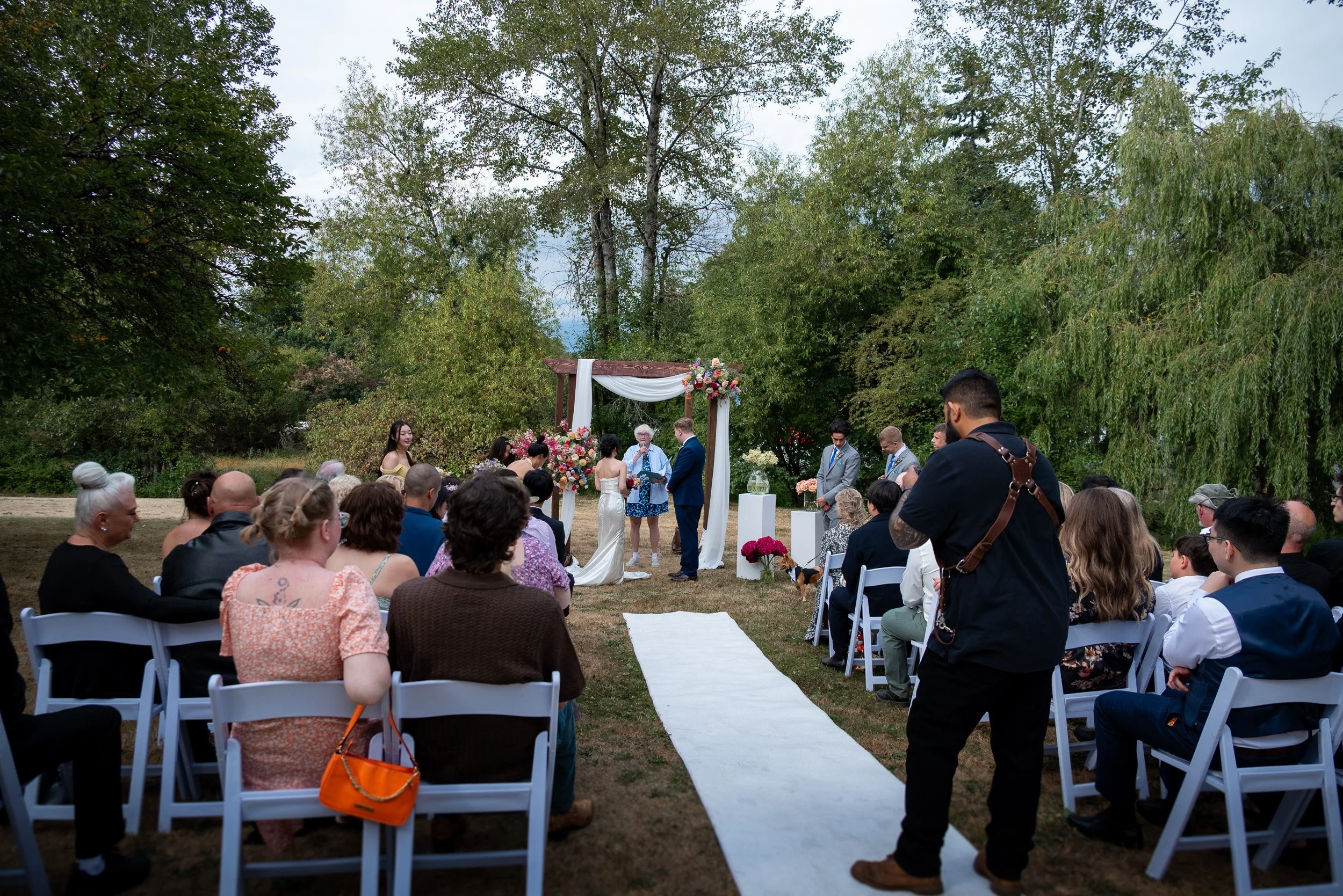 Outdoor wedding ceremony with guests seated on white chairs, a white aisle runner, floral arrangements, and a wedding officiant speaking in front of a decorated arch. Tall trees and overcast sky in the background.