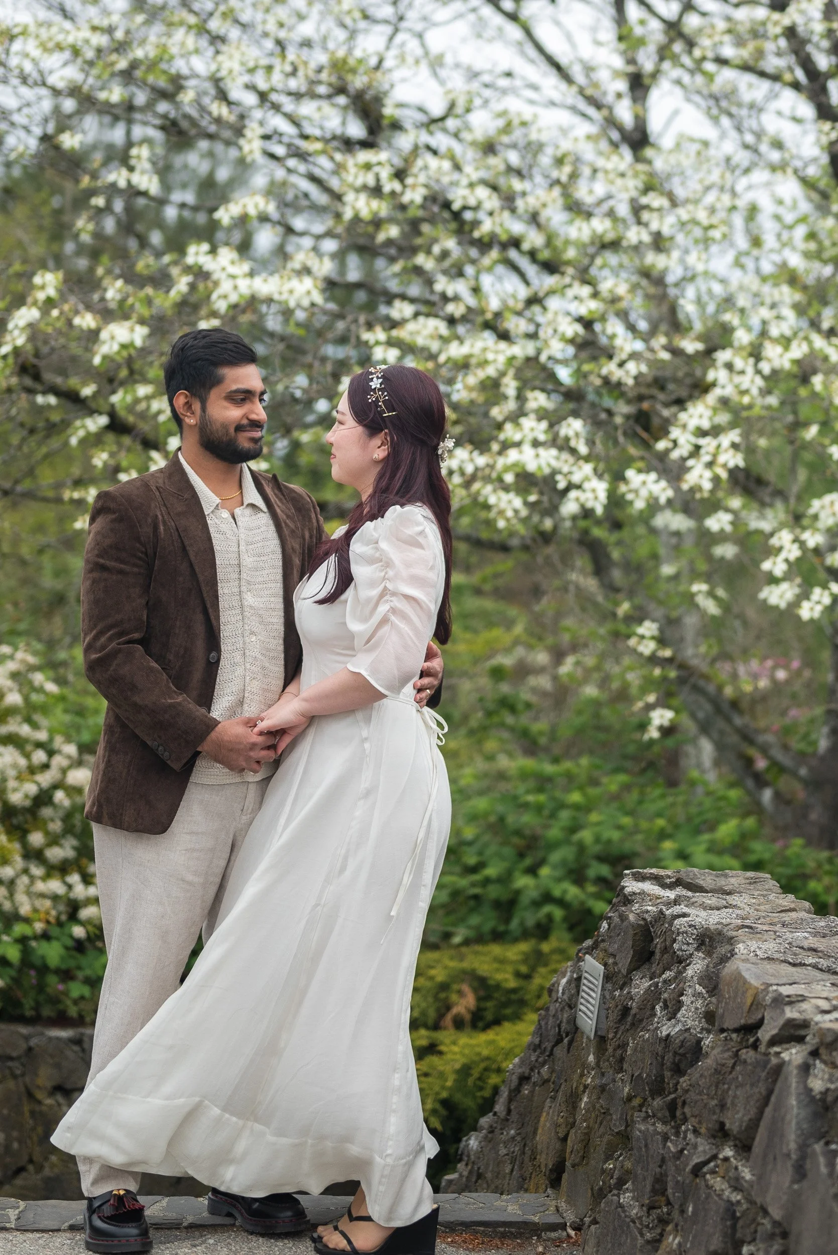 A newlywed couple gazing into each other's eyes outdoors, standing on a stone path with cherry blossom tree in the background. This is a wedding at Queen Elizabeth Park with Cherry blossoms.