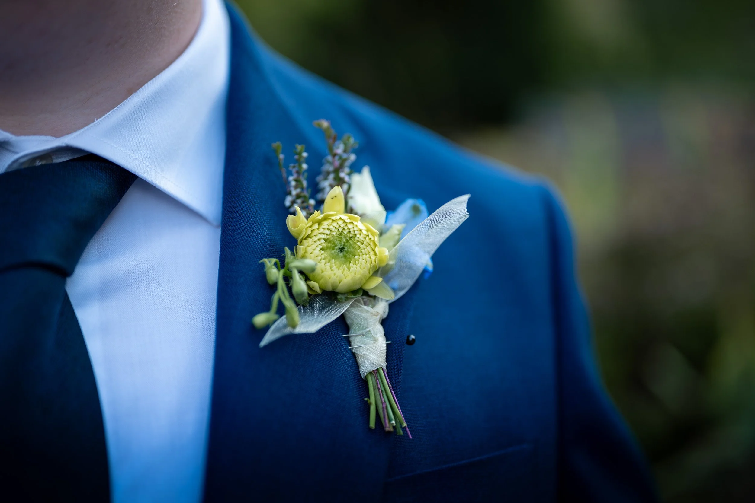 Close-up of a groom's upper body wearing a blue suit and white shirt, with a boutonniere made of yellow and white flowers pinned to the lapel, outdoors.