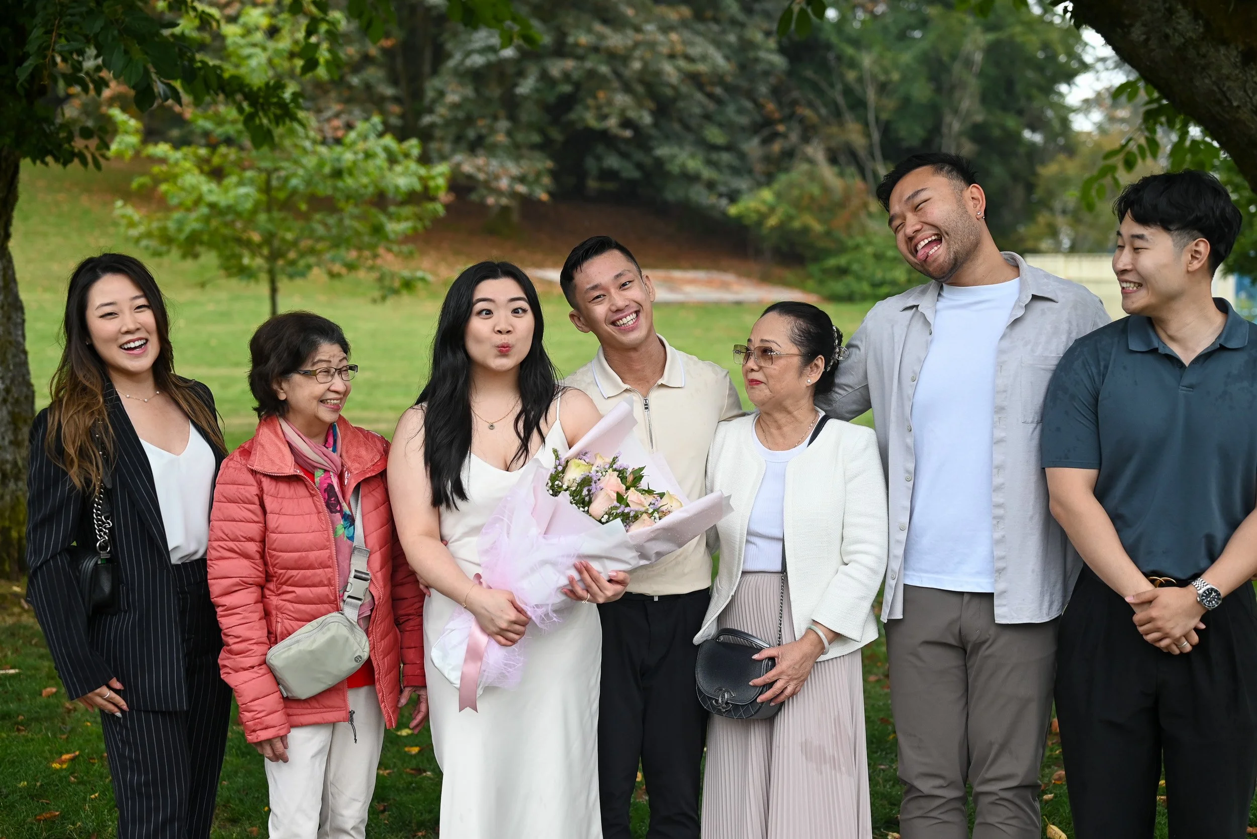 A diverse group of nine people standing outdoors under a tree, smiling and celebrating, with one woman holding a bouquet of flowers.