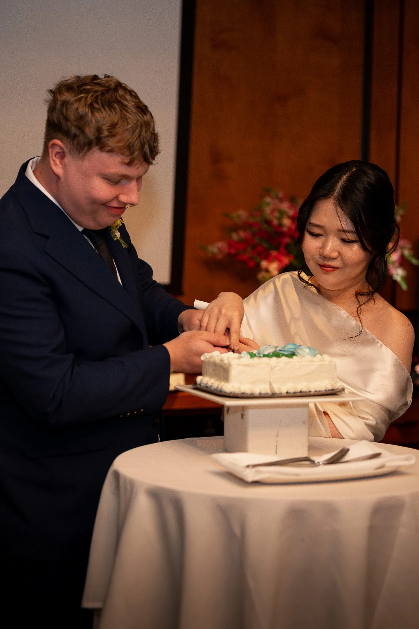 A man and woman in formal attire participate in a wedding ceremony, cutting a wedding cake together. The man is wearing a dark suit, and the woman is in a cream satin dress. There are flowers in the background.