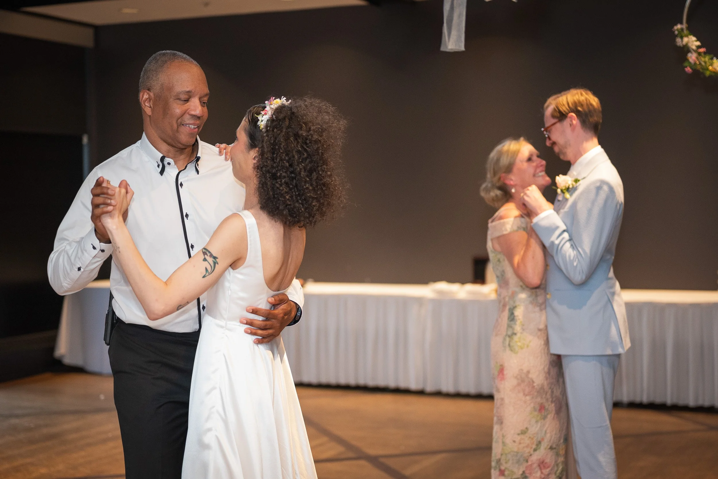 Two couples dancing at a wedding reception. The couple in the foreground is dressed in formal attire, with the woman in a white dress with a floral headband and the man in a white shirt and black pants. The woman has tattoos on her arm and is smiling