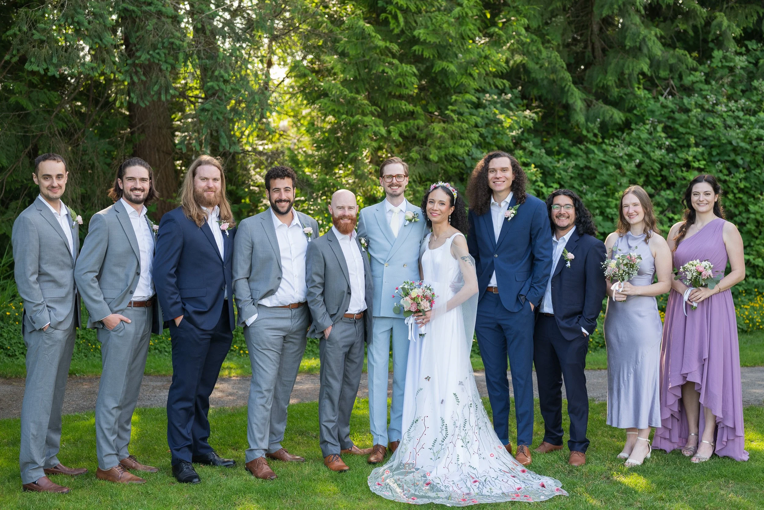 A wedding party with 11 people standing outdoors in front of trees, dressed in formal attire with some holding bouquets, smiling for a group photo.