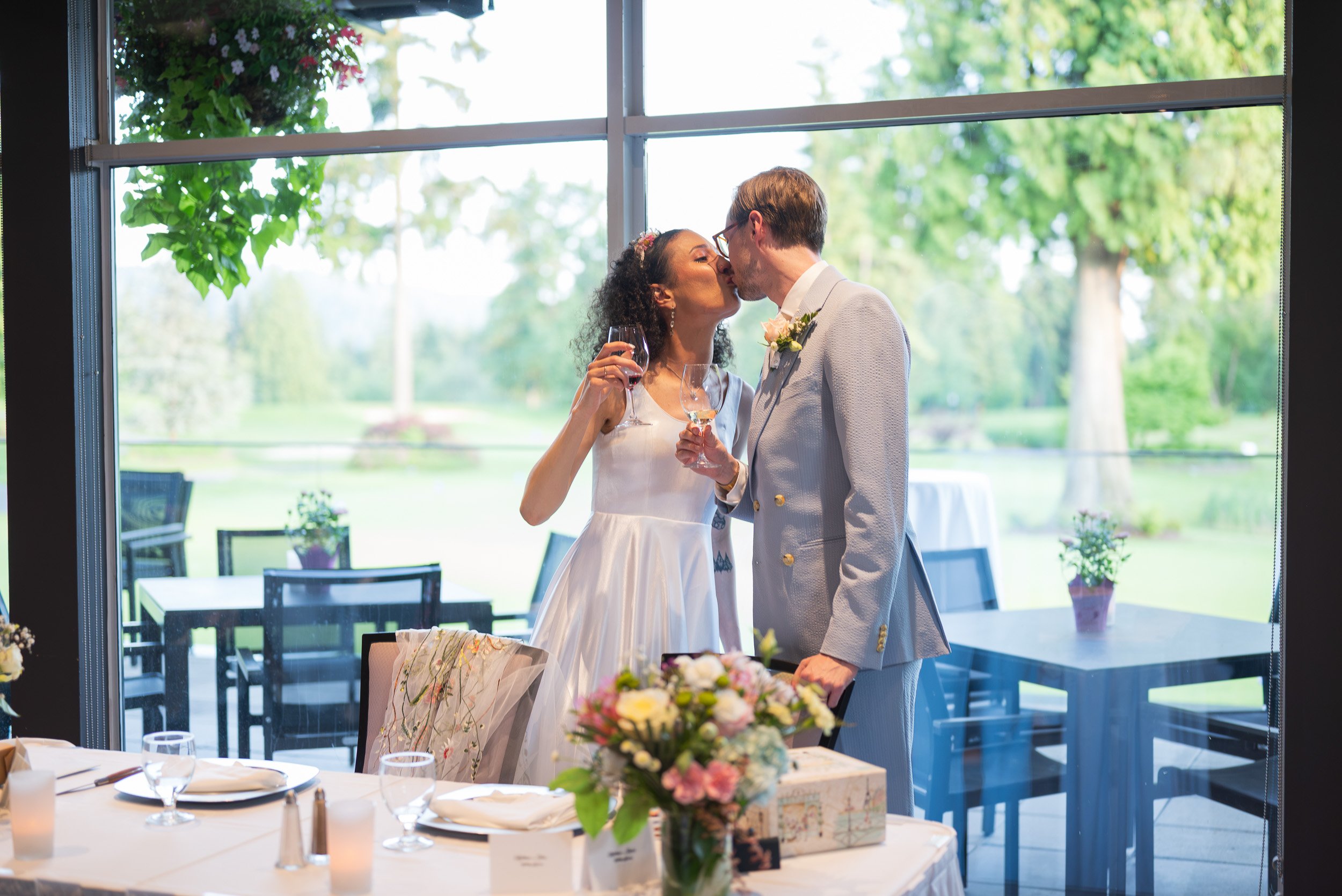 A bride and groom kiss during their wedding reception at Syemour Golf and Country Club, with a large window showing green trees outside. The bride holds a glass of red wine, and there are flowers and table settings in the foreground.