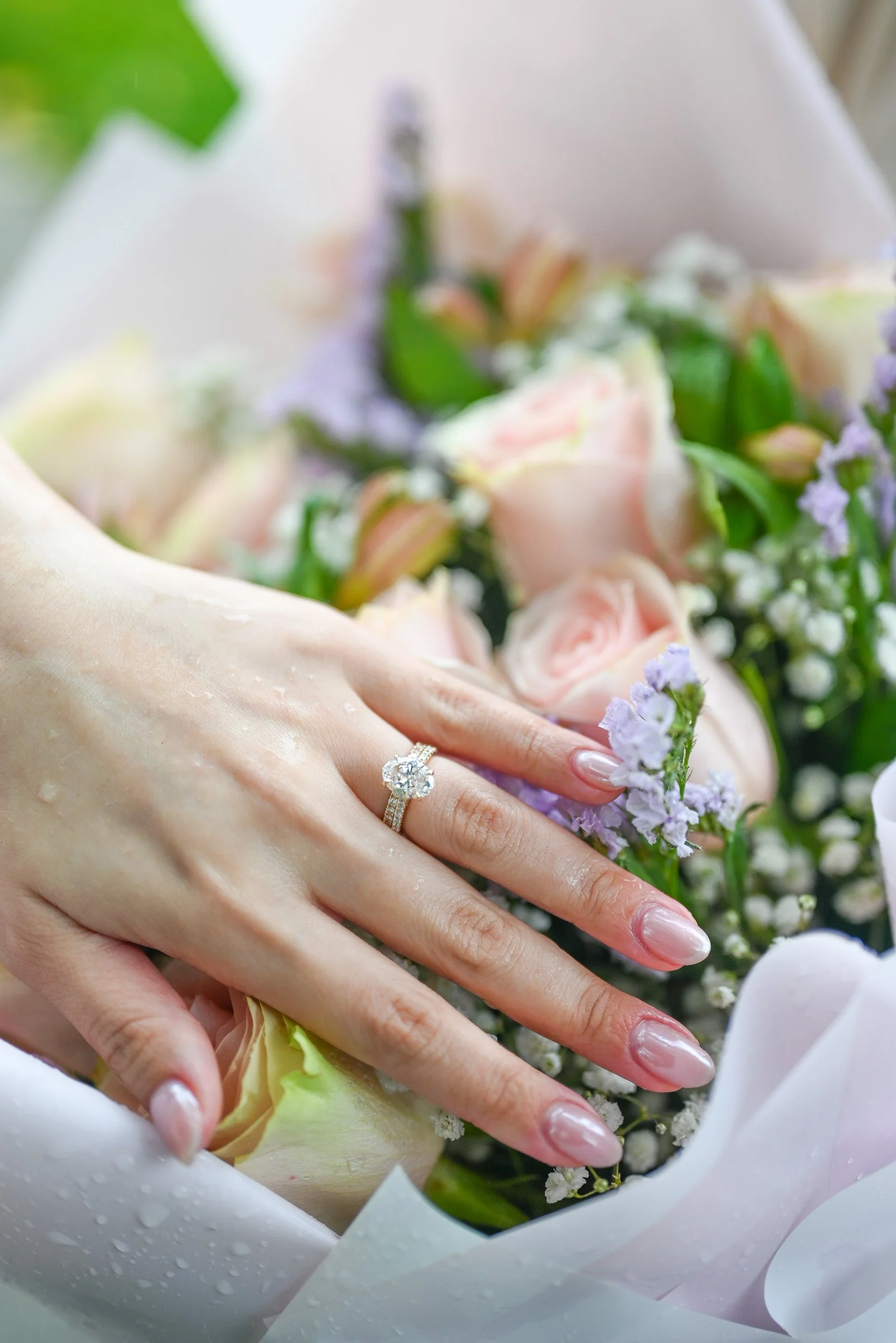 Close-up of hands with wedding rings on a bride's fingers, surrounded by a bouquet of pink roses, white orchids, and purple flowers wrapped in white paper with water droplets on it.