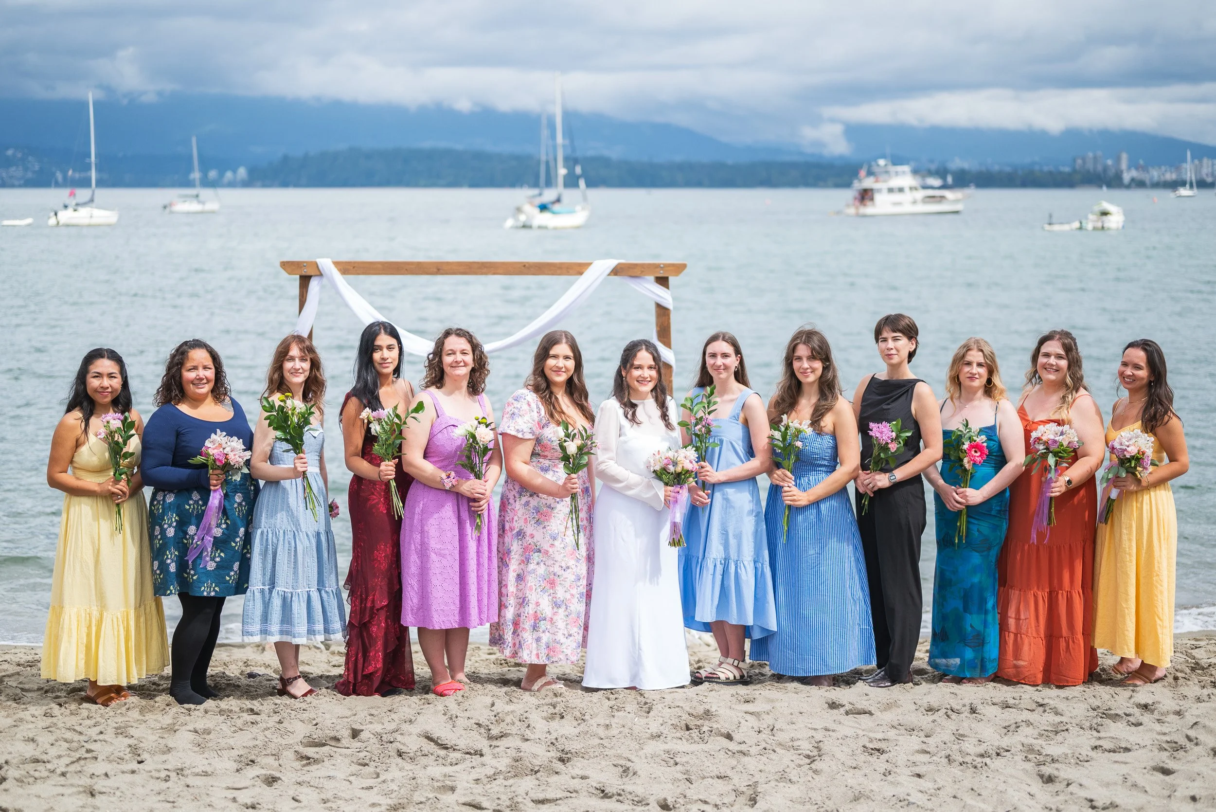 A group of women in colorful dresses standing on a sandy beach, holding bouquets, with sailboats on the water and mountains with cloudy sky in the background.