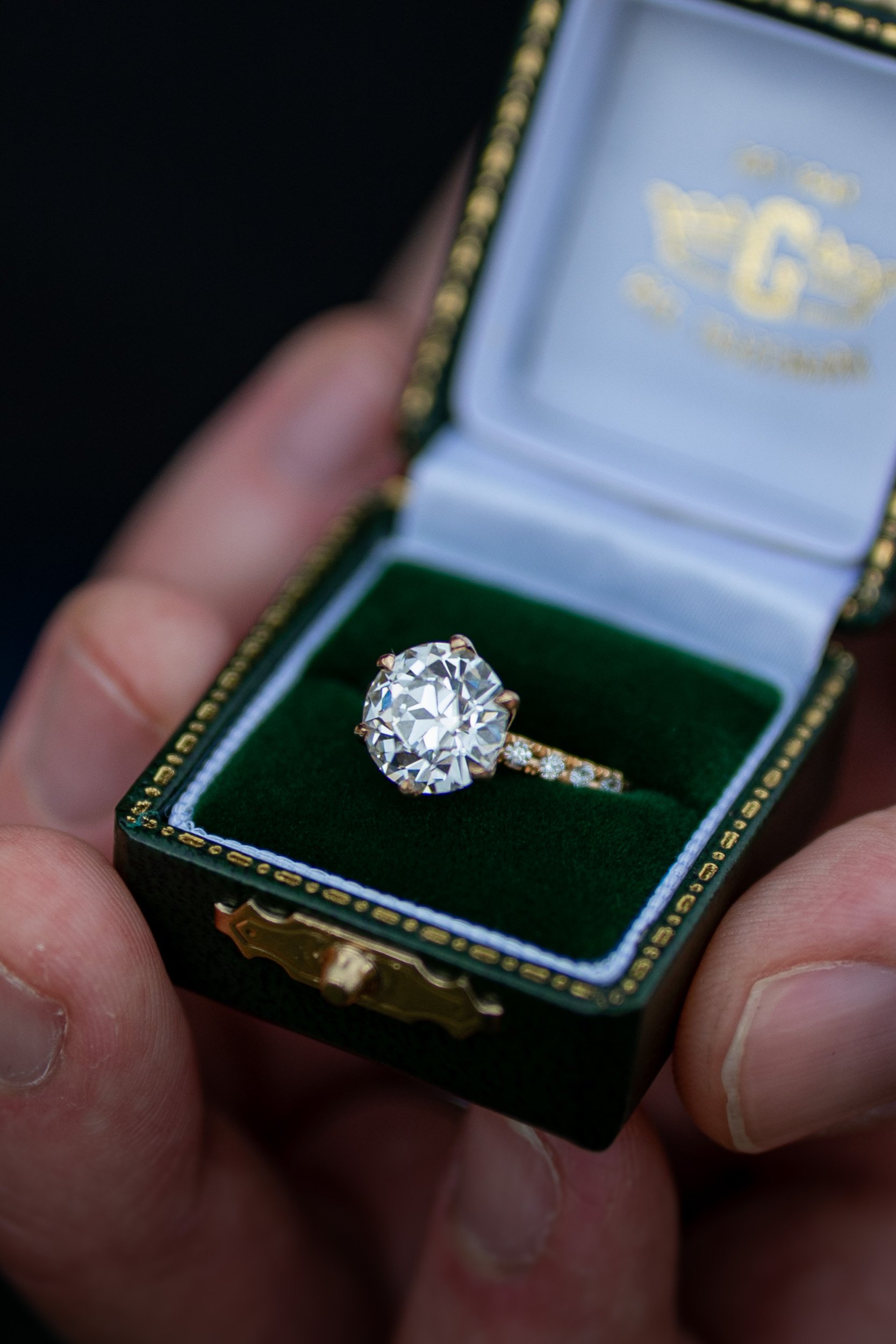Close-up of a diamond engagement ring with a large oval-cut diamond in a gold setting, displayed in an open jewelry box with a green velvet interior.