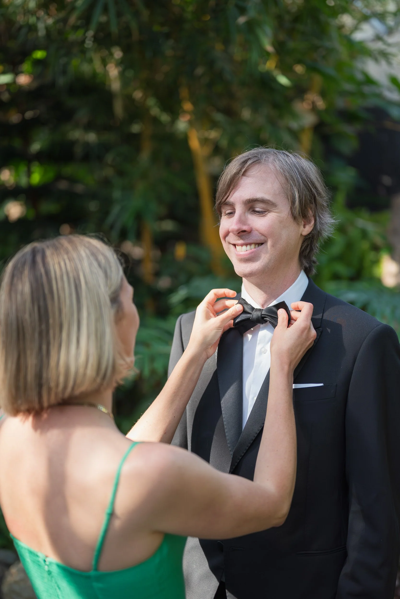 A man in a tuxedo and bow tie smiling while a woman in a green dress adjusts his bow tie. They are outdoors with lush green foliage in the background.