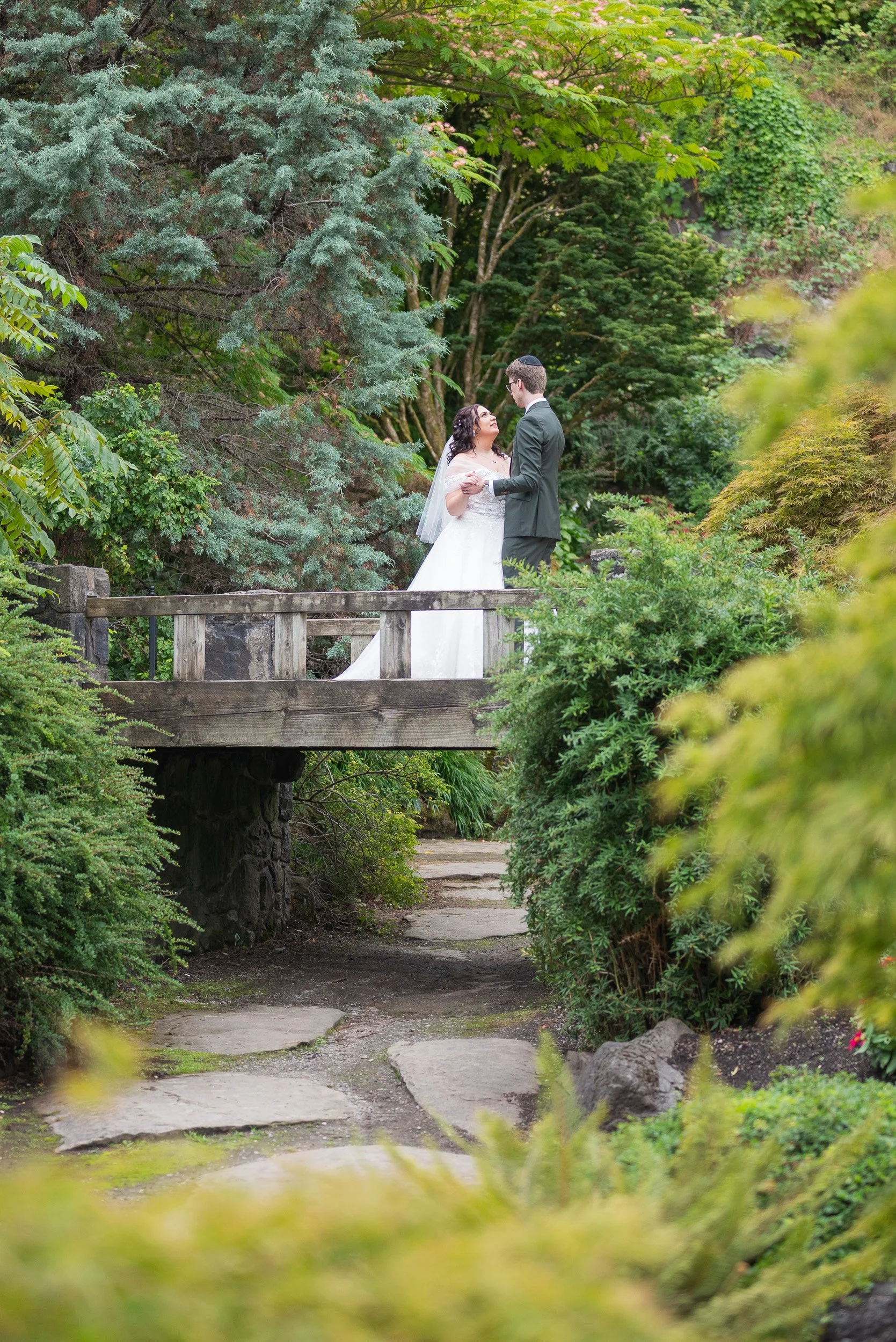 A couple dressed in wedding attire standing on a small bridge surrounded by lush greenery, trees, and bushes at Queen Elizabeth Park Wedding.