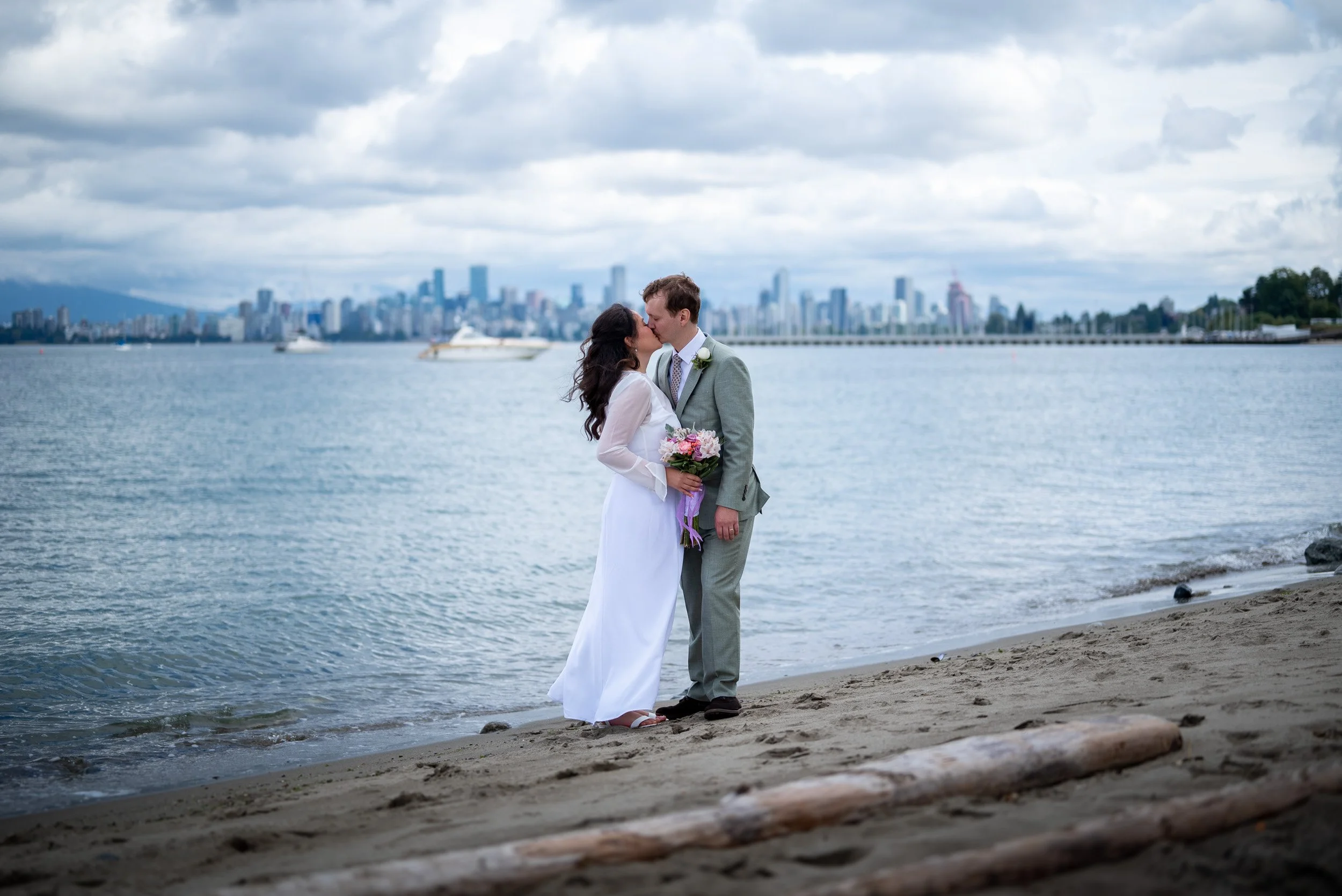 A bride and groom kiss on a beach at Jericho Beach with a Vancouver city skyline and cloudy sky in the background. The bride holds a bouquet of flowers, and they are dressed in wedding attire.