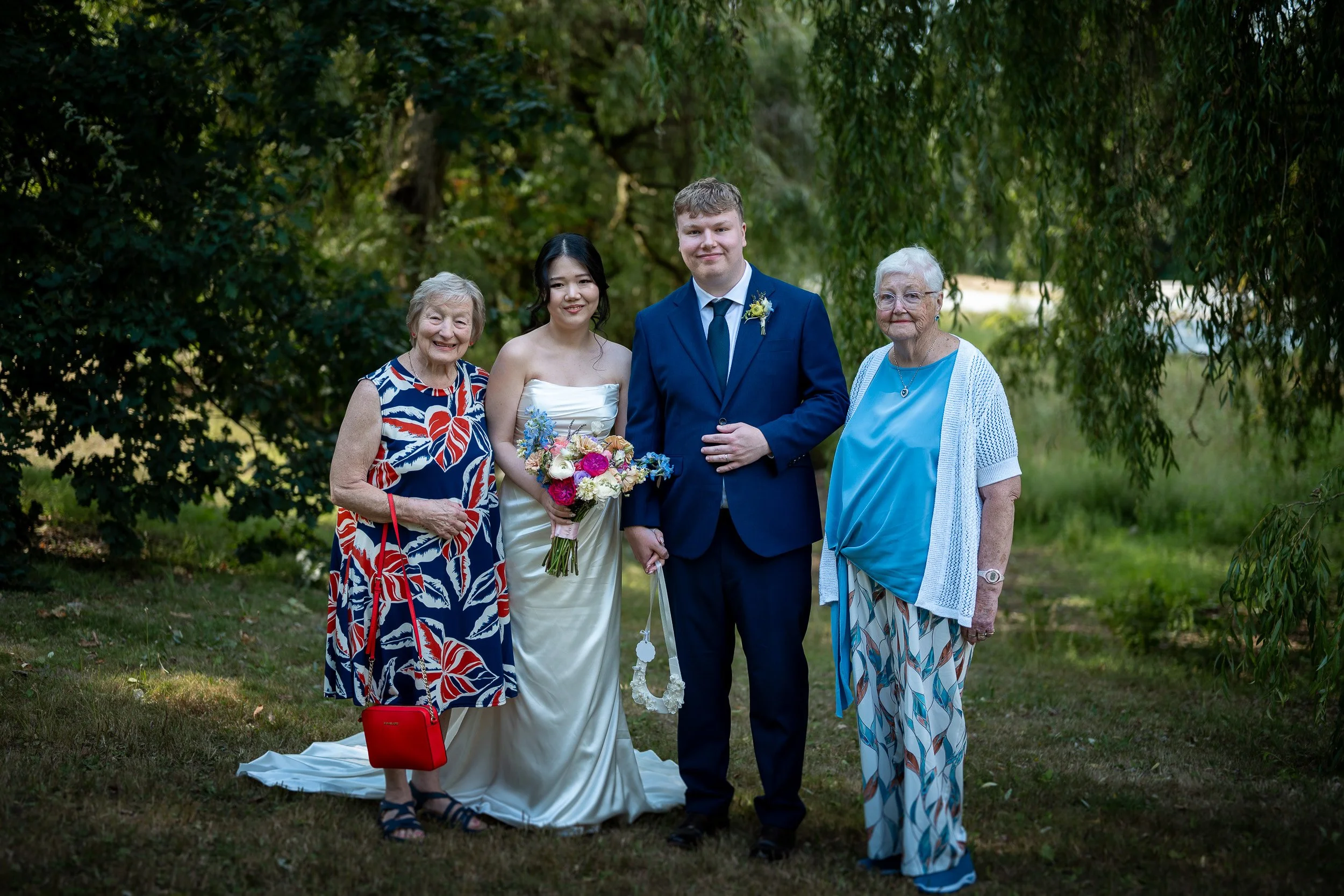 A bride in a white wedding dress holding a bouquet stands with three elderly women outdoors; the bride is flanked by two women on her left and right, in casual summer dresses. The group is standing on grass in a park-like setting with trees in the ba