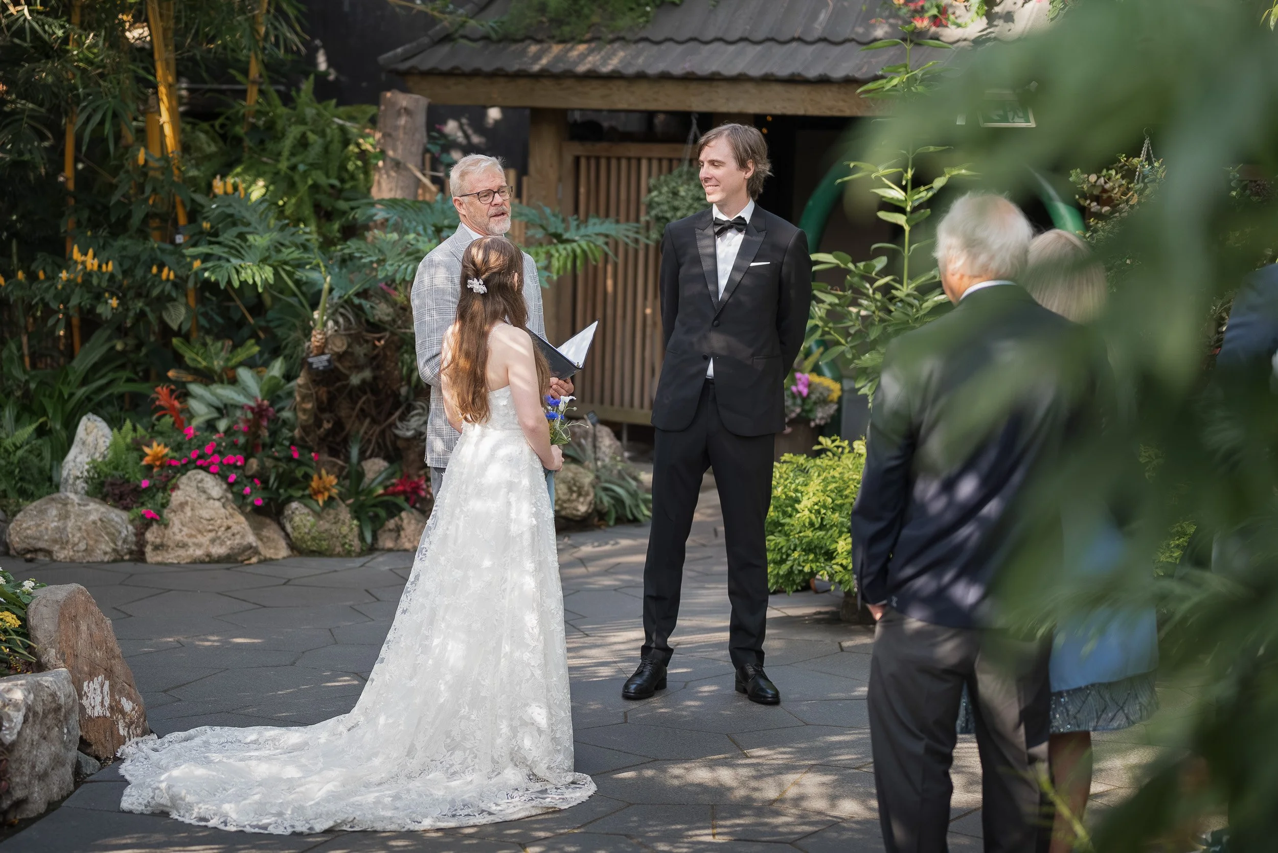A wedding ceremony taking place at Bloedel Conservatory with a bride, groom, and officiant surrounded by lush greenery, flowers, and rocks.
