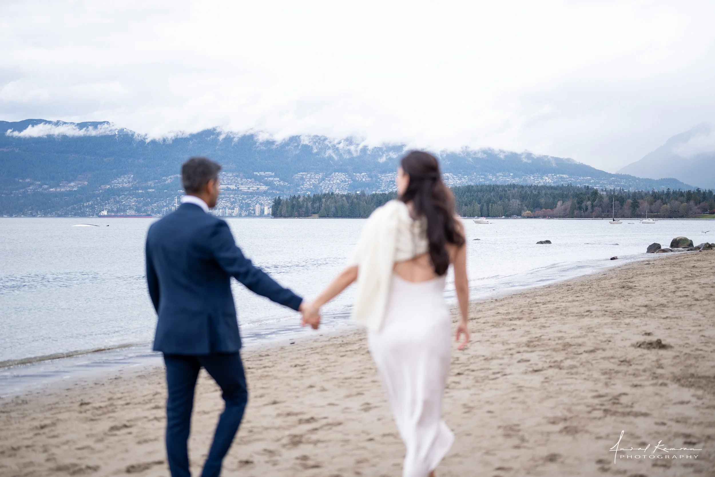 A couple holding hands and walking on the beach with mountains, trees, and boats in the background, overcast sky.