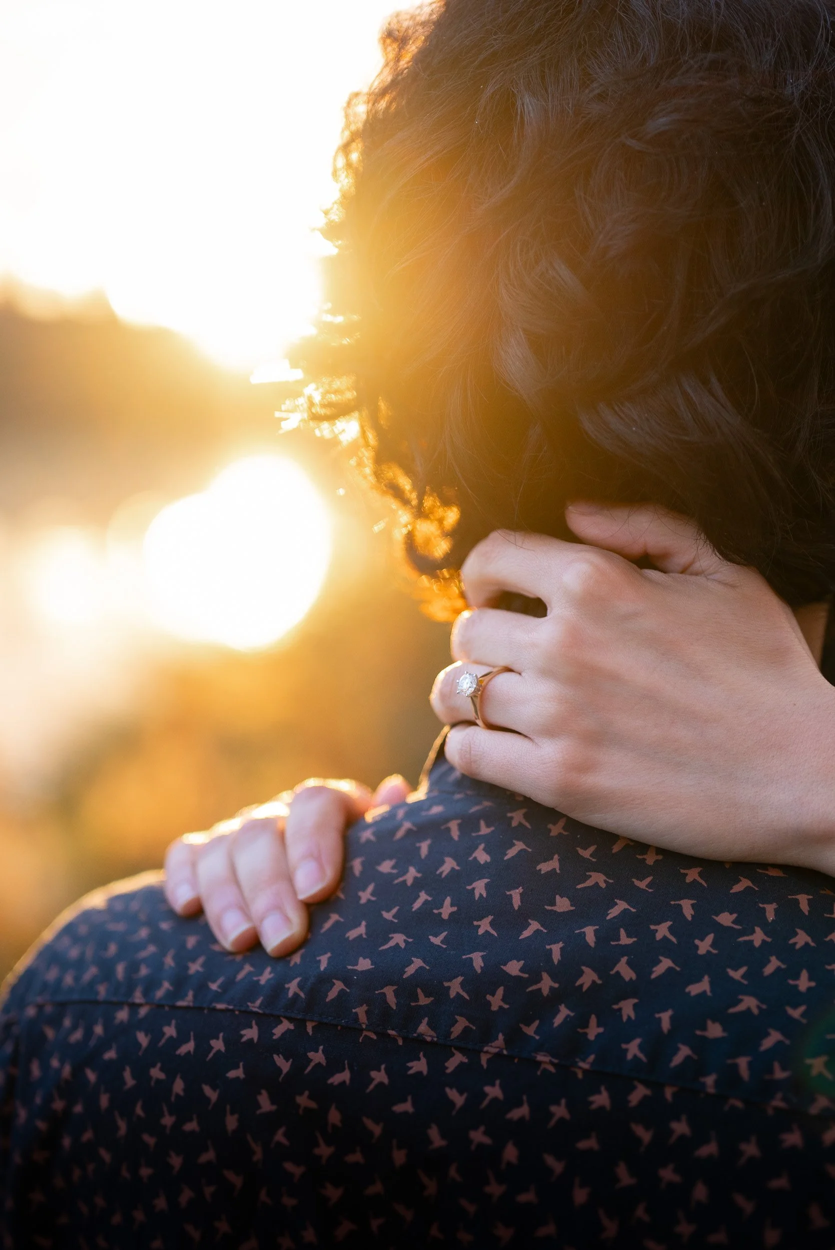 Close-up of two people hugging during sunset, with the focus on their arms and hands, one person wearing a ring.