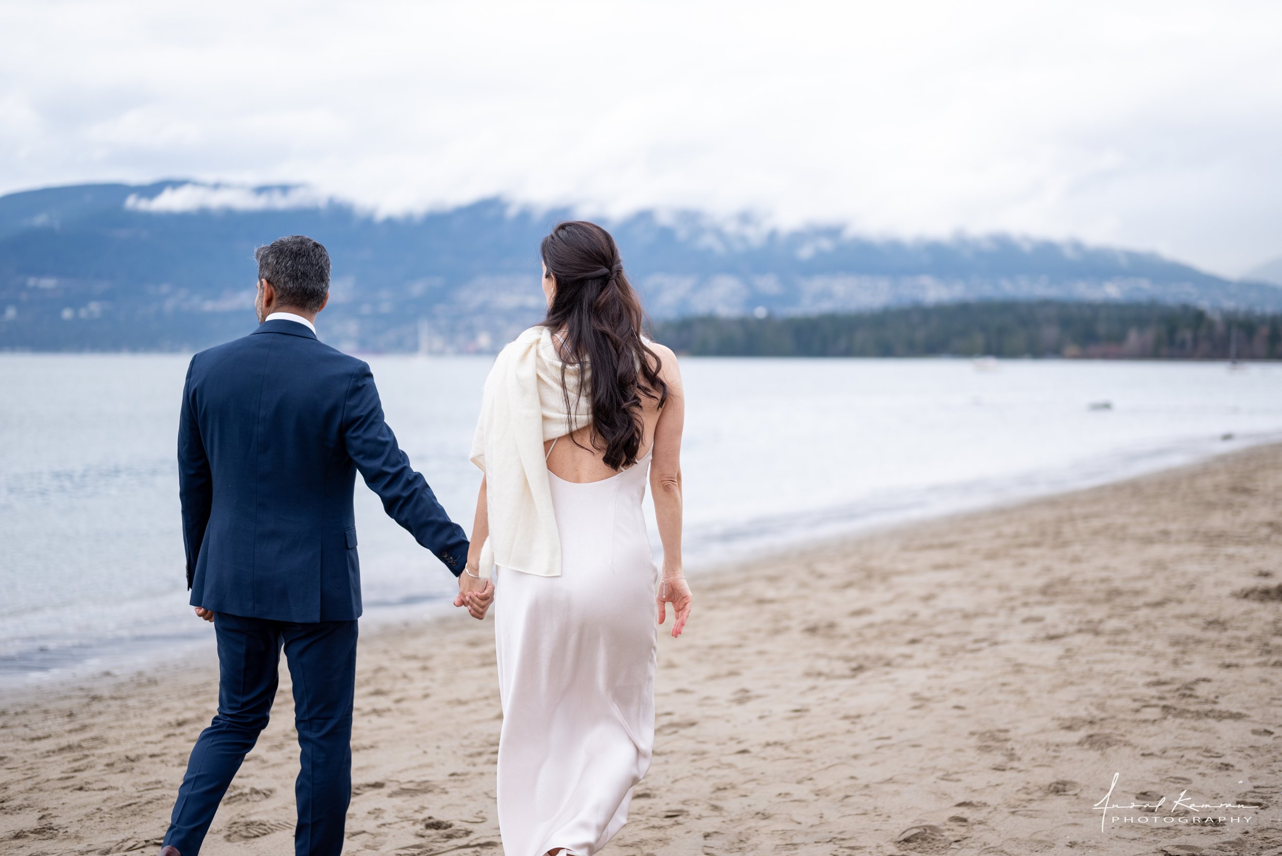 A newly-wed couple in wedding attire walking hand in hand on a beach with mountains in the background at Kitsilano Beach in Vancouver