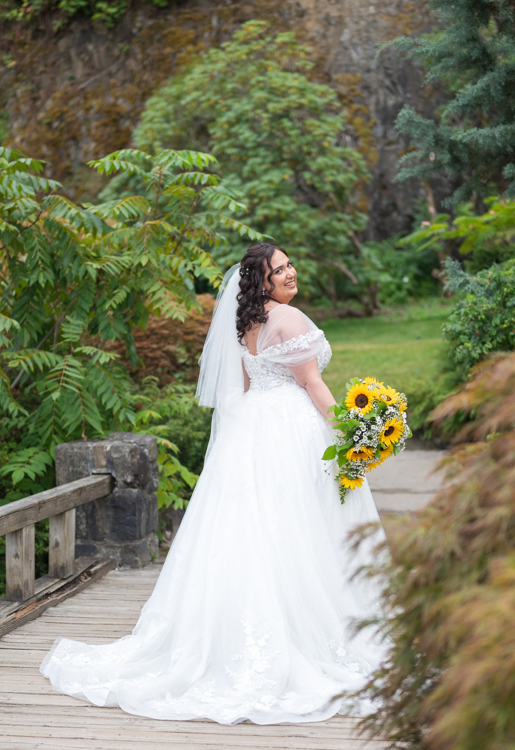 A bride in a white wedding dress with lace details, holding a bouquet of sunflowers and white flowers, standing on a wooden bridge in a lush green garden in Queen Elizabeth Park, Vancouver, BC.