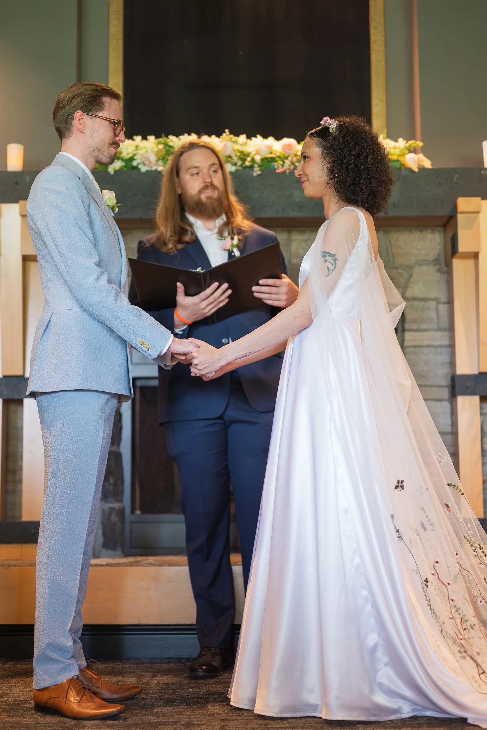 A couple getting married during a wedding ceremony, holding hands and facing each other, with an officiant standing behind them, in an indoor setting decorated with flowers and candles at Seymour Golf and Country Club.