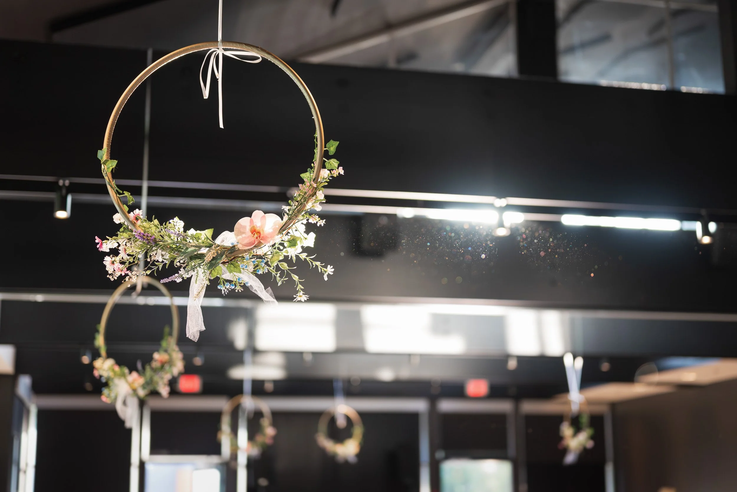 Hanging floral wreath with pink and white flowers is suspended from the ceiling in a modern interior space with black ceiling panels and hanging light fixtures.
