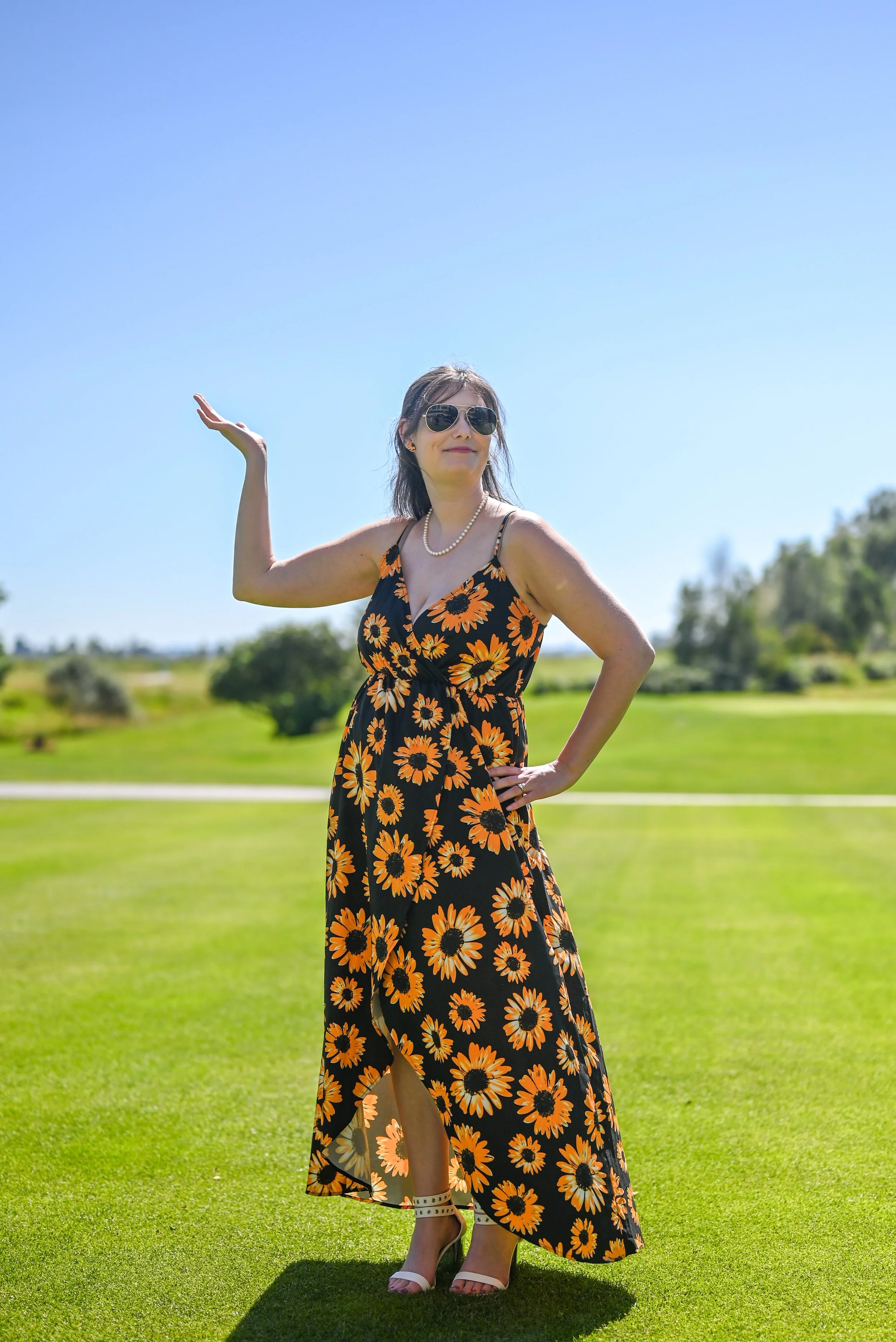Woman in a sunflower-patterned dress standing on grass under a clear blue sky, wearing sunglasses, pearl necklace, and high heels, with her hand raised in a gesture.